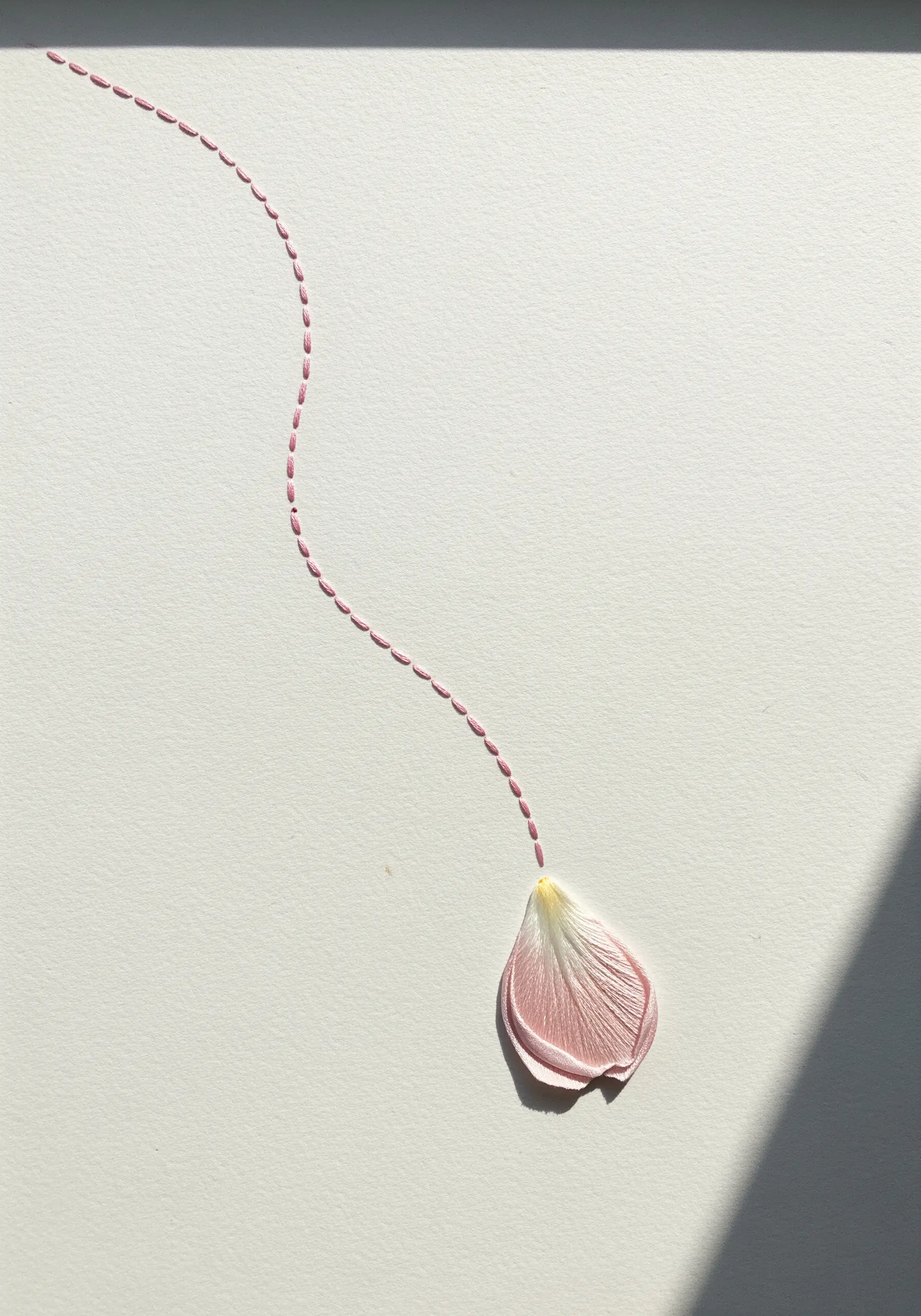 A single embroidered petal at the end of a curved, dashed line of pink stitches on white paper.