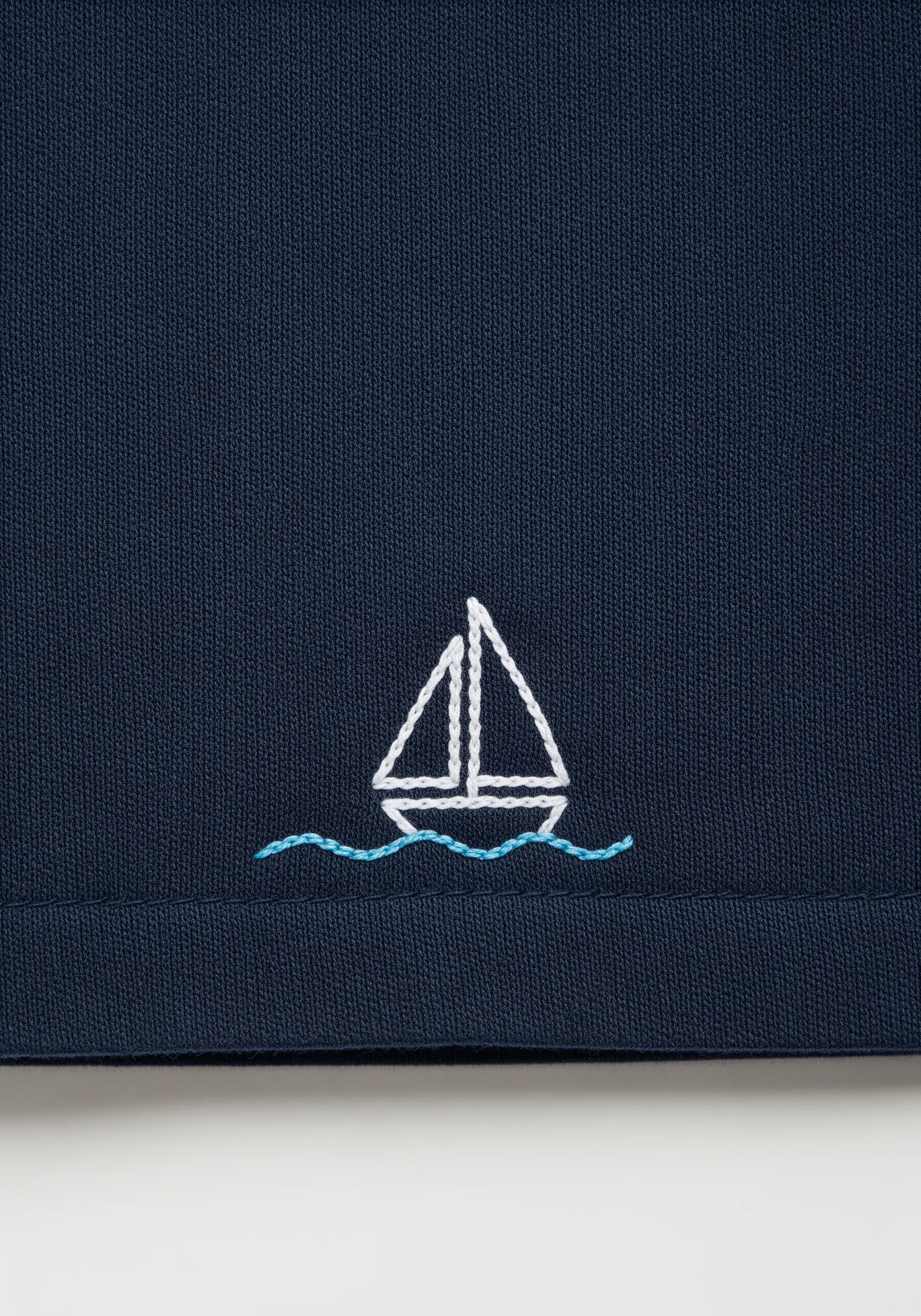A tiny, minimalist sailboat embroidered in white and blue on dark navy fabric.