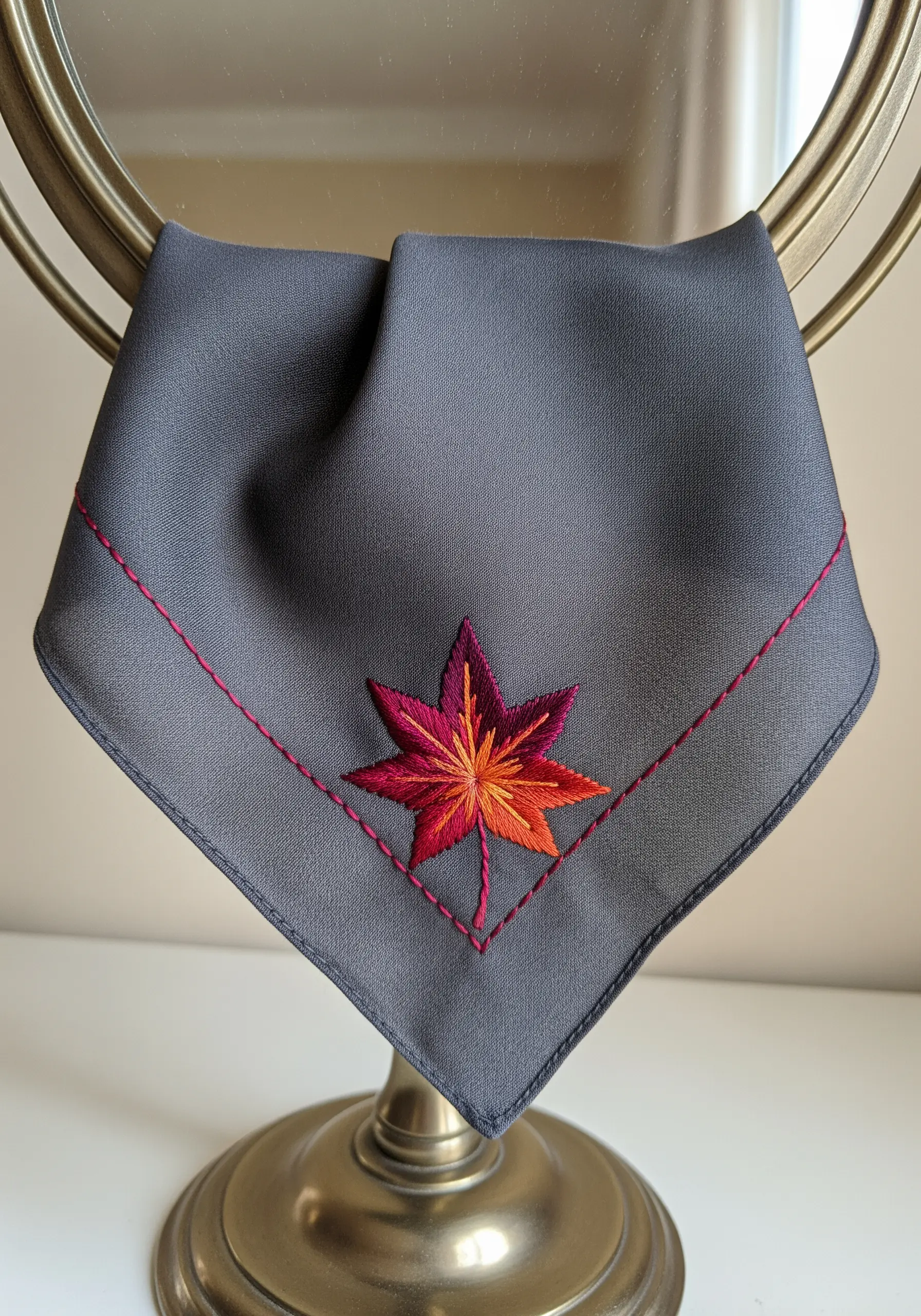 A single maple leaf embroidered with a color gradient from yellow to red on a gray handkerchief.