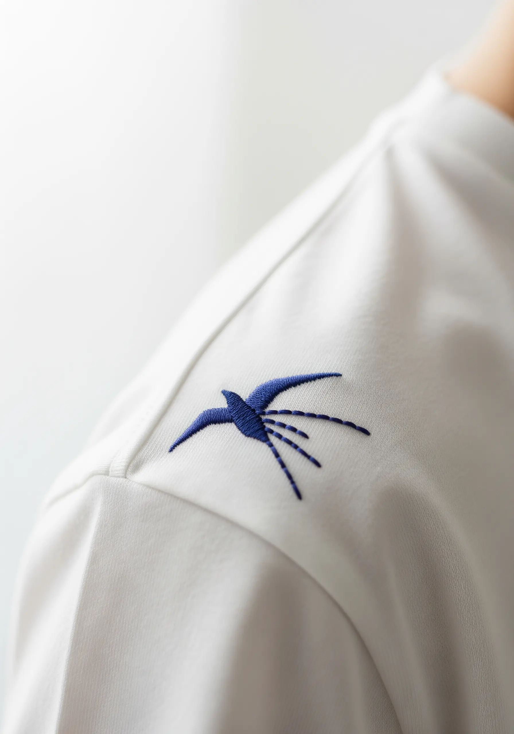 A stylized blue swallow bird embroidered on the shoulder of a t-shirt.
