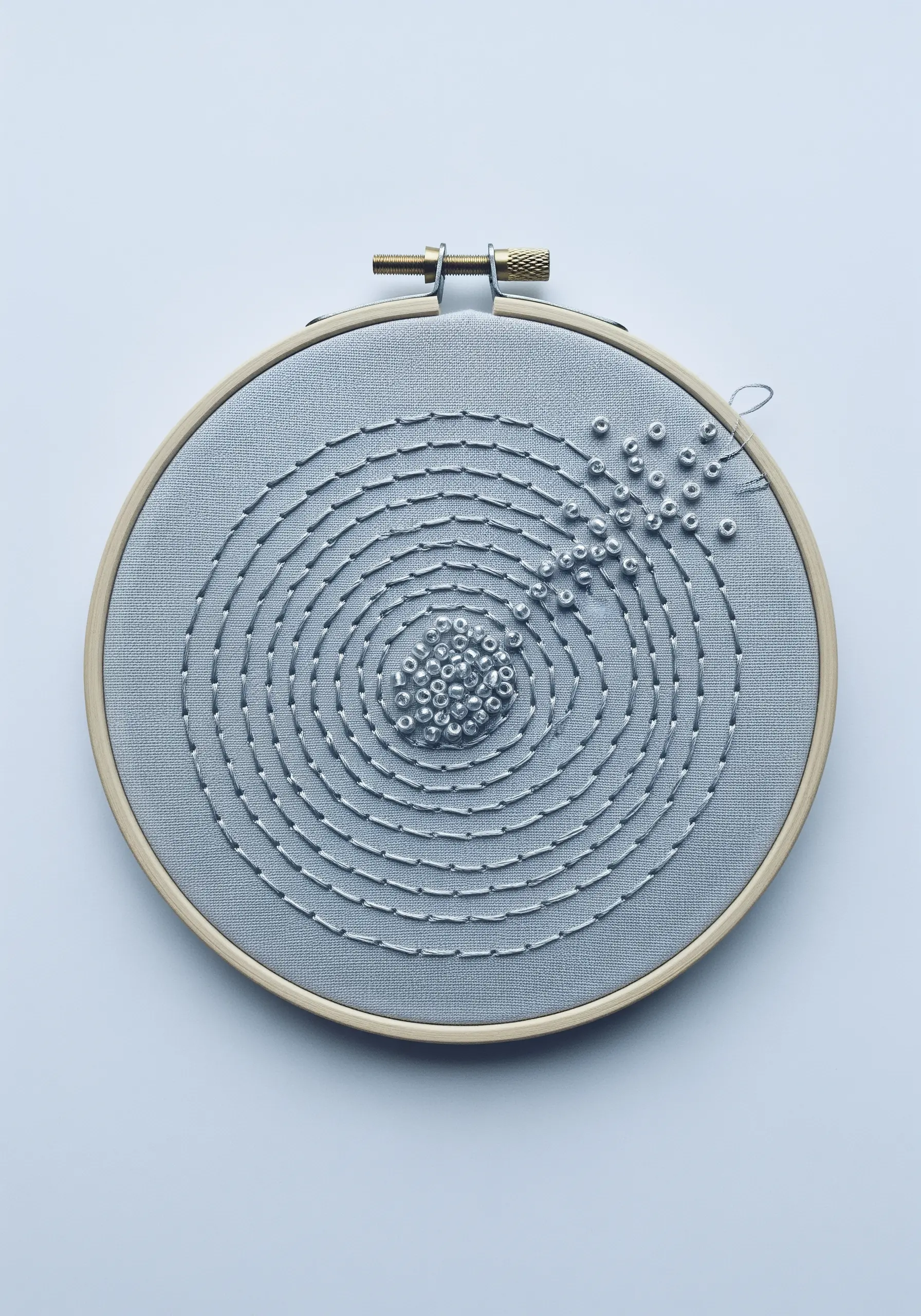 Minimalist hoop art showing a spiral of silver beads on gray fabric, unraveling at the edge.