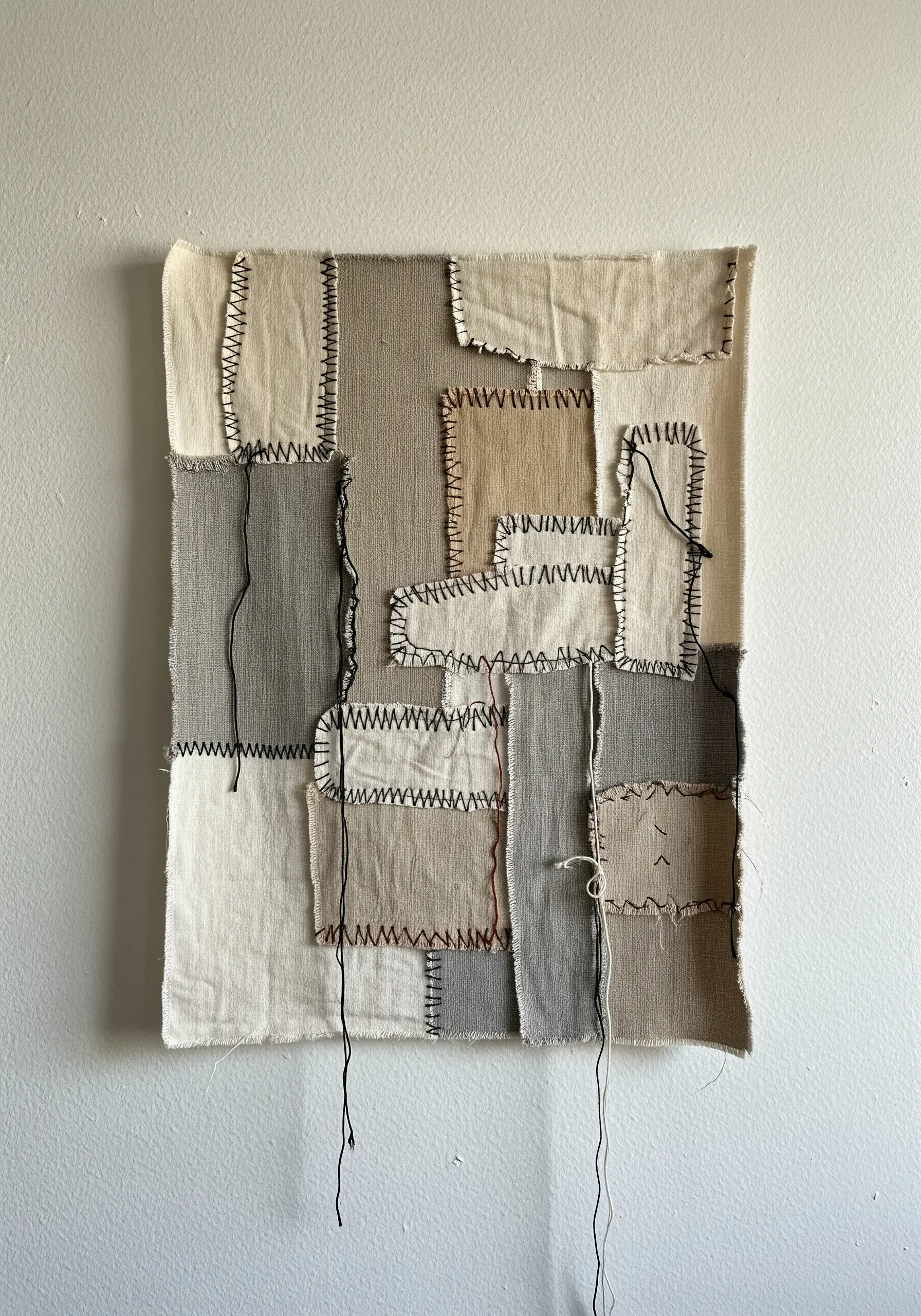 A neutral-toned abstract patchwork with frayed edges and thick, visible black stitching.