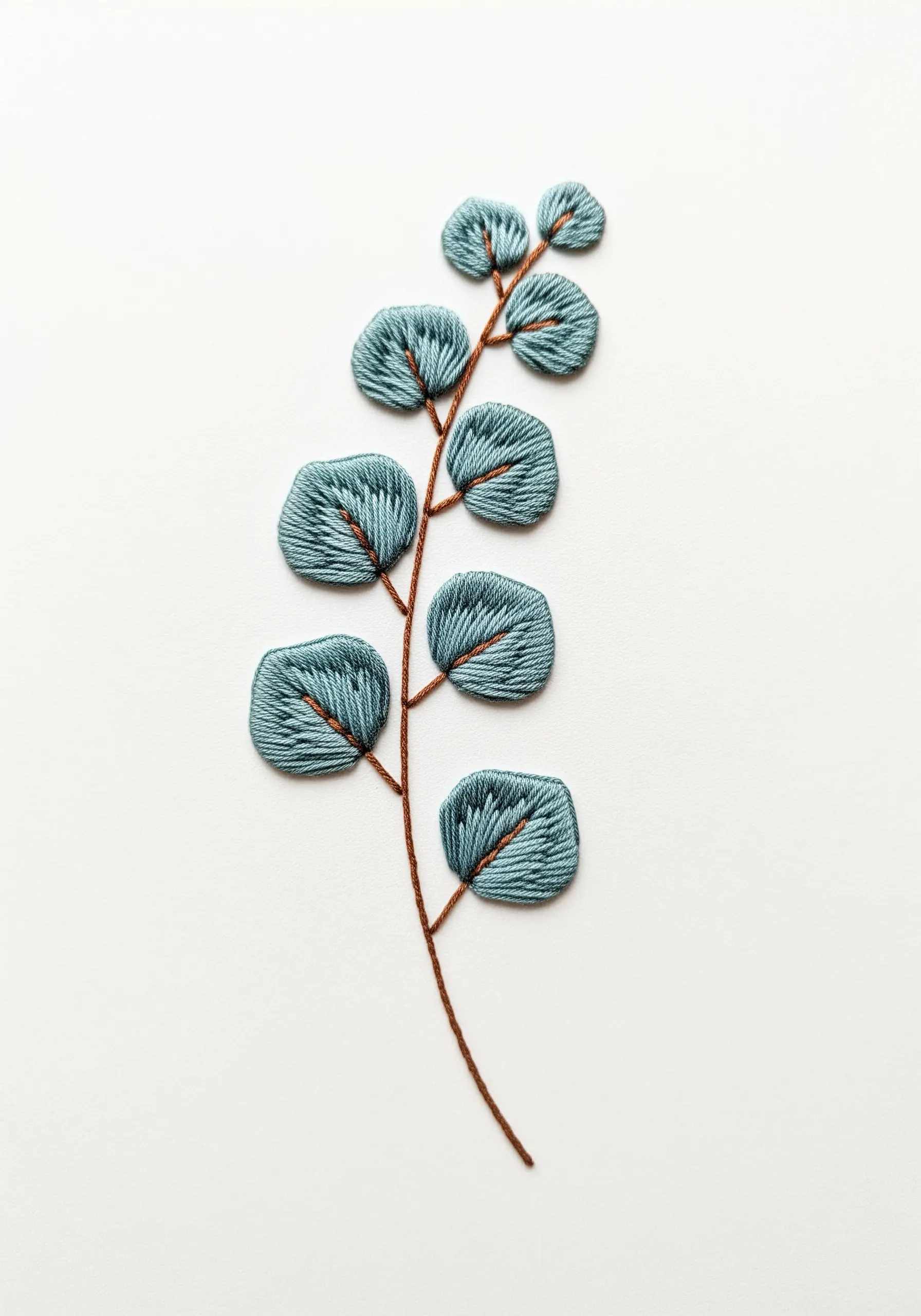 A sprig of eucalyptus embroidered with muted teal satin stitch on white fabric.