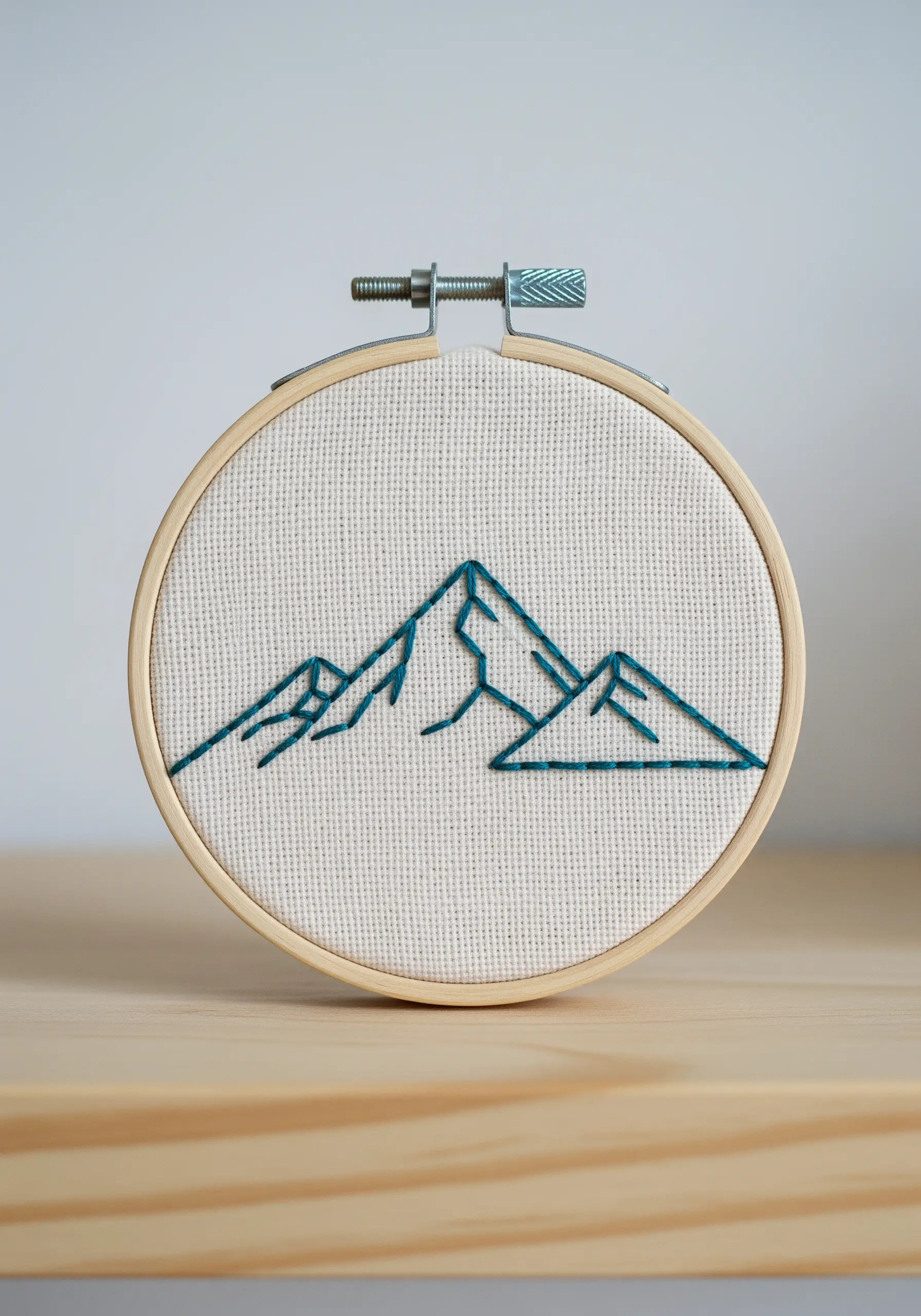 Minimalist mountain range outline stitched in dark teal thread on neutral fabric.