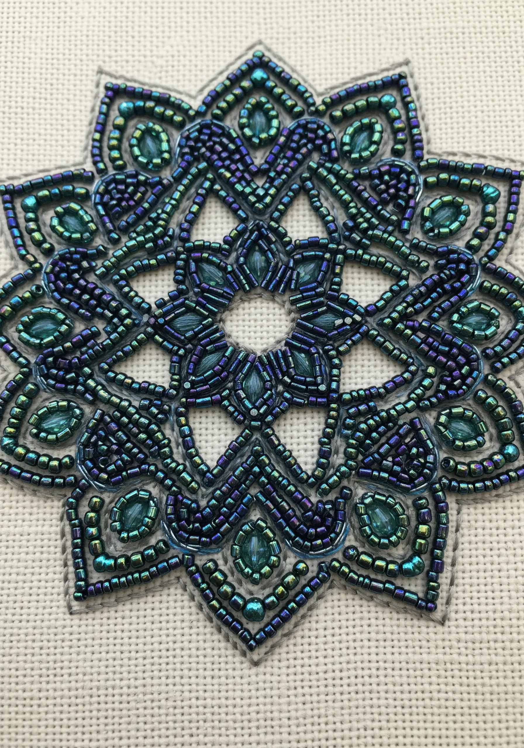 A close-up of an intricate star-shaped mandala created entirely with iridescent blue and green seed beads.