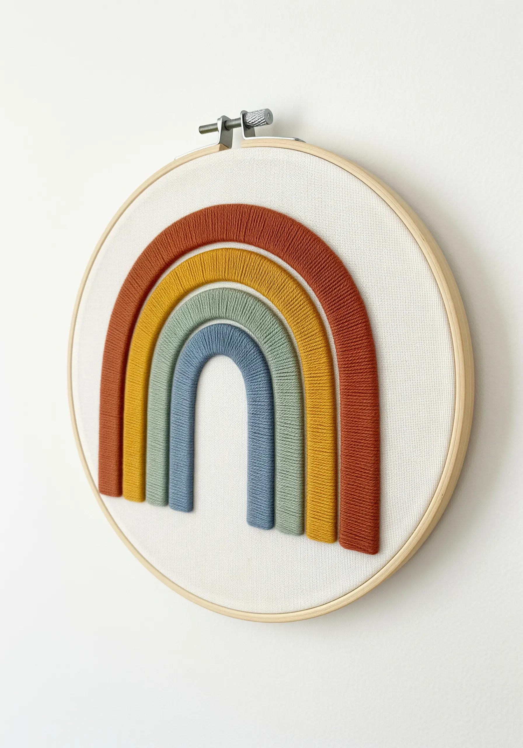 A modern embroidery hoop with a raised rainbow made from wrapped yarn in earthy tones.
