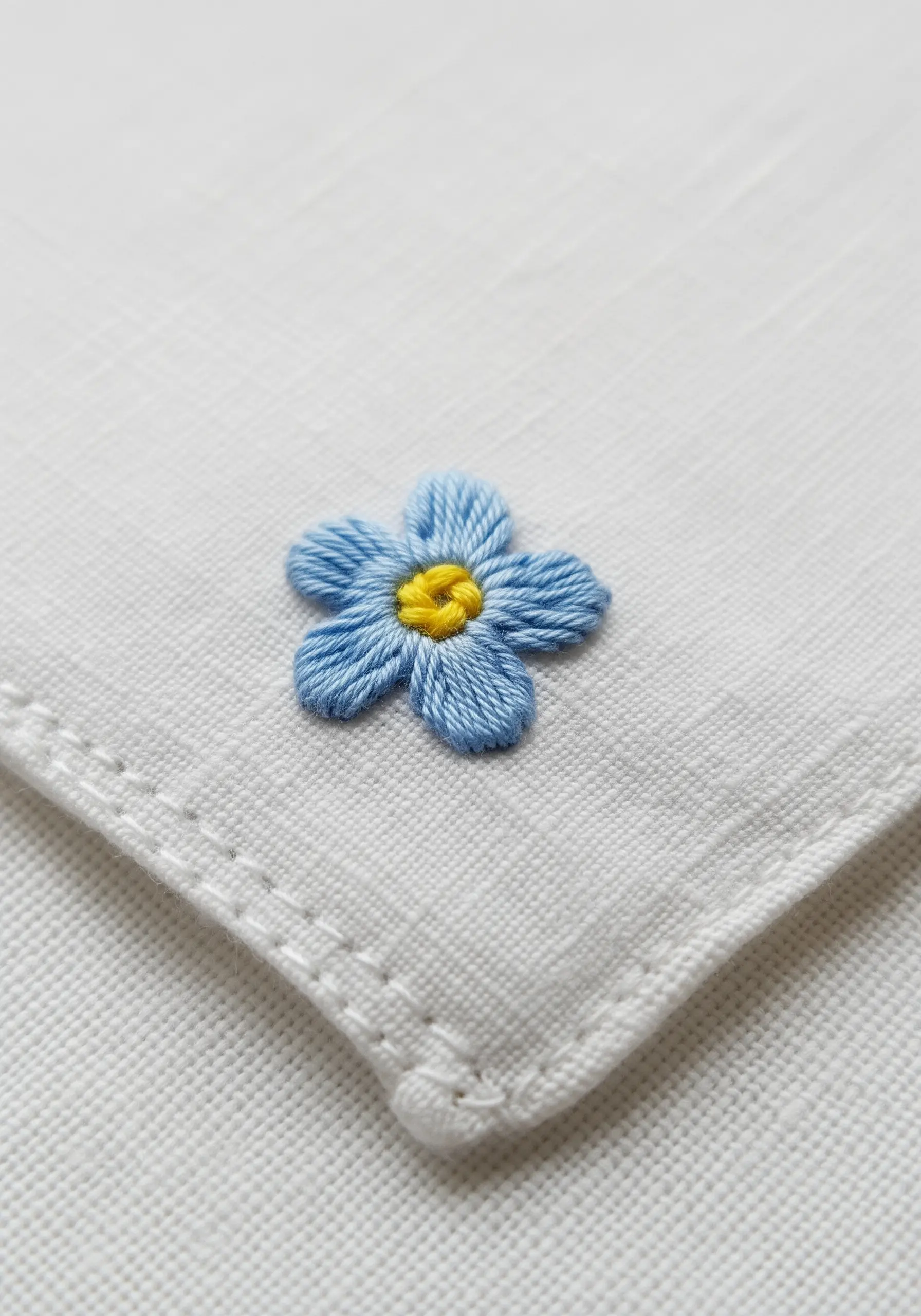 A tiny, blue forget-me-not flower stitched with lazy daisy petals on a white handkerchief.