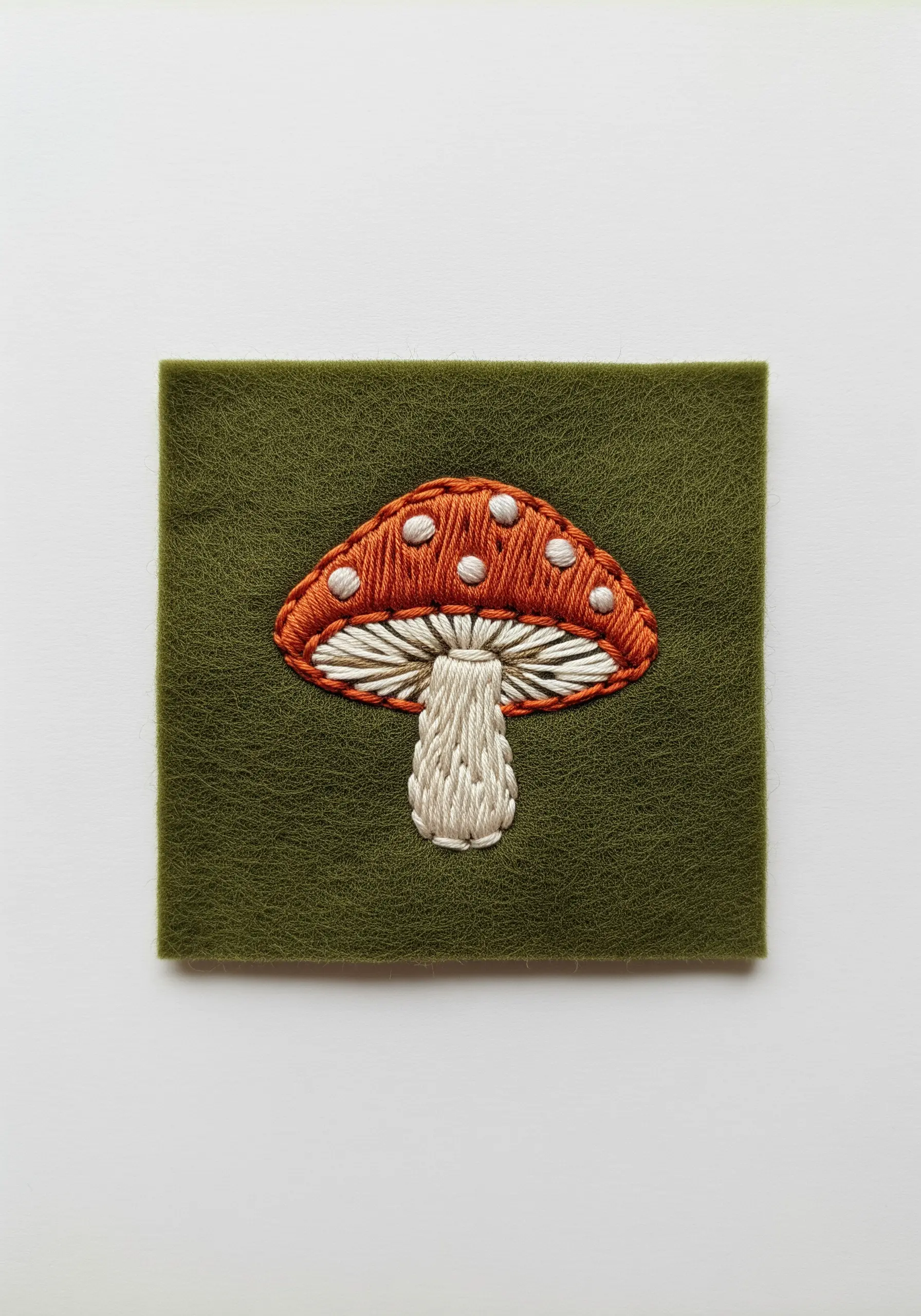 A realistic embroidered mushroom with a red cap and white spots on green felt.