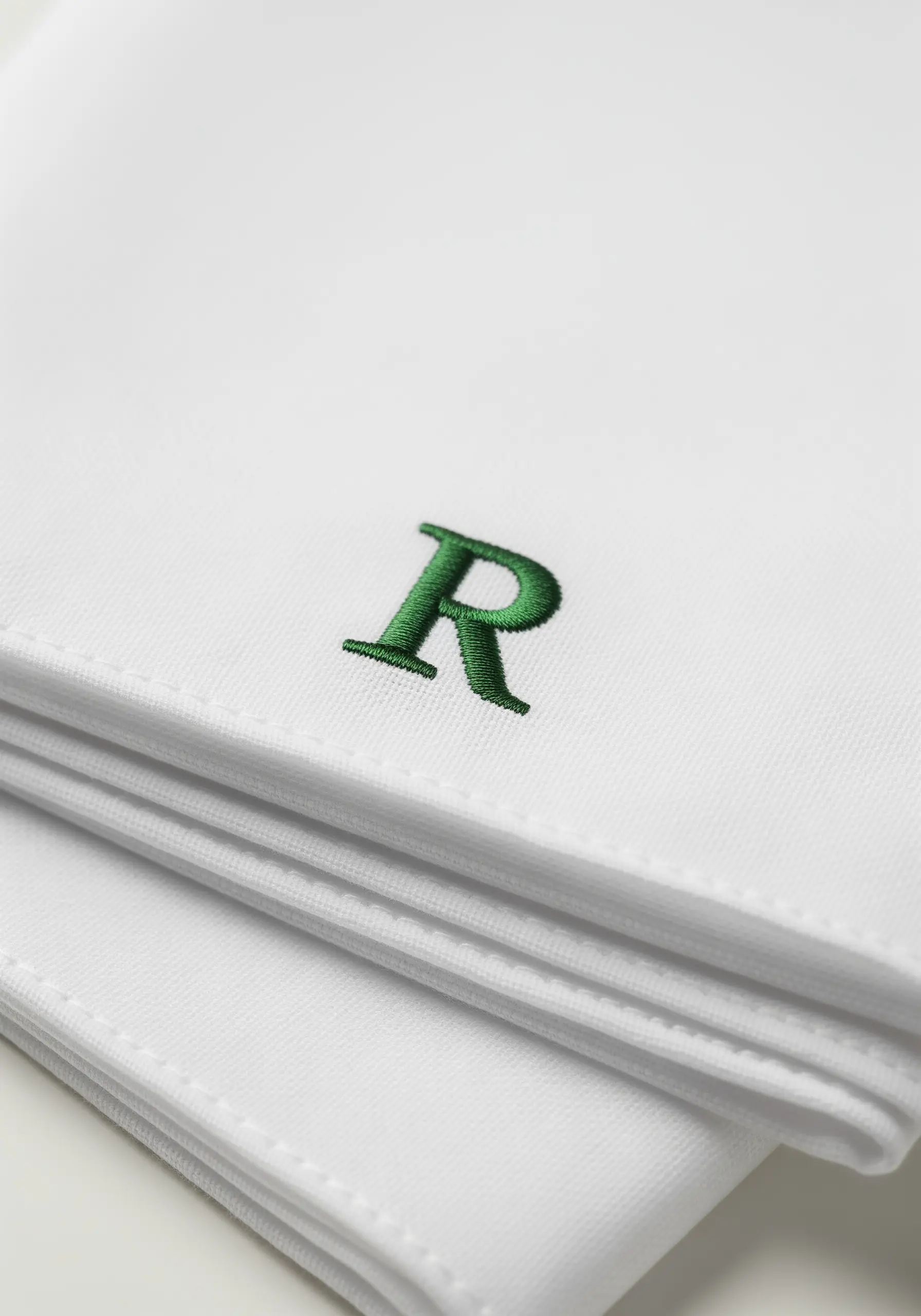 A crisp, dark green serif monogram 'R' stitched on a stack of folded white handkerchiefs.