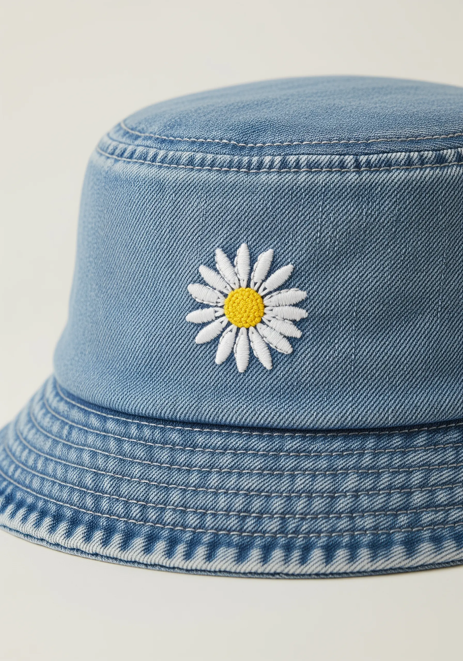 A single white and yellow daisy embroidered on the front of a light blue denim bucket hat.