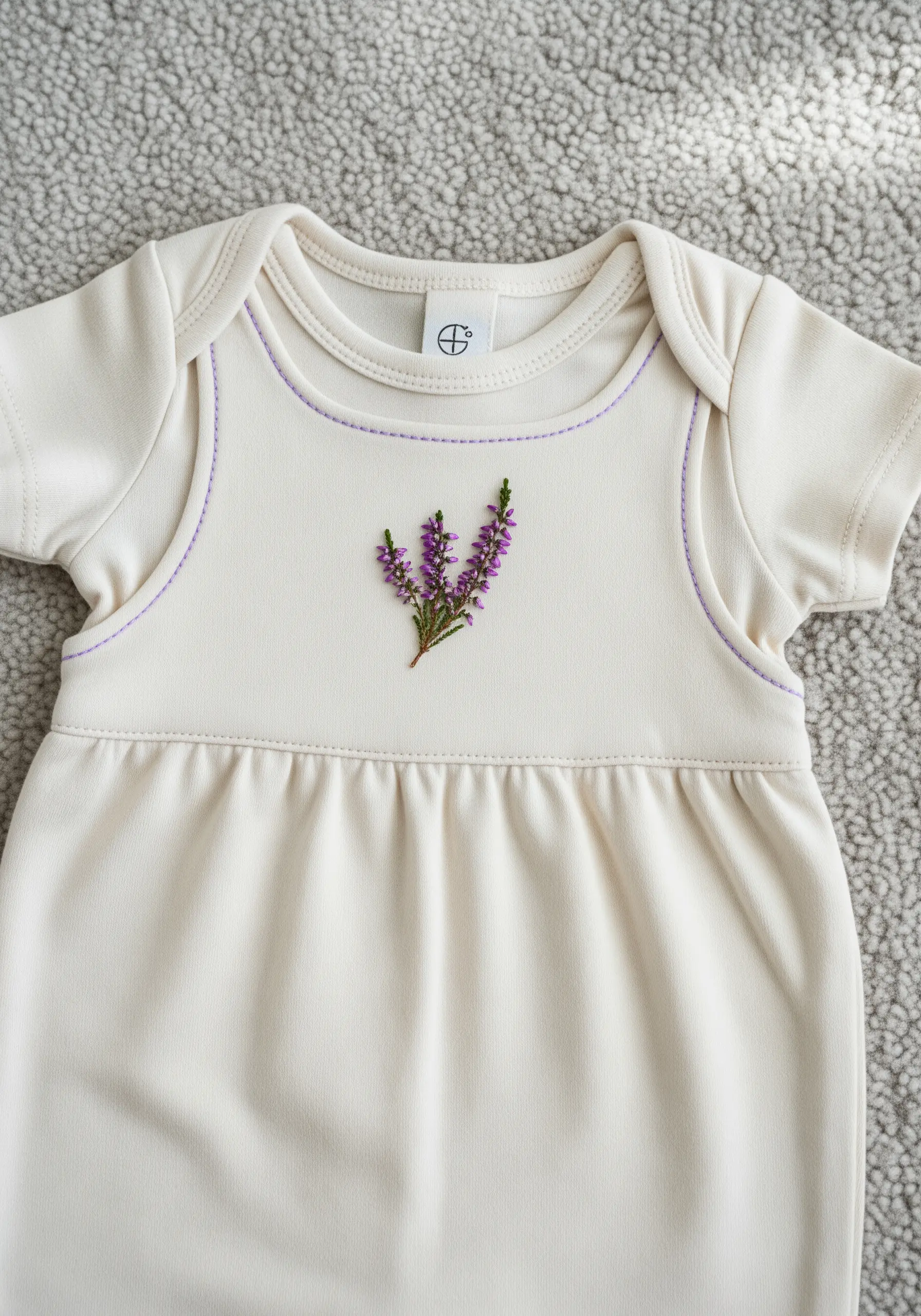 A delicate sprig of pressed heather embroidered on the chest of a baby's cream-colored gown.