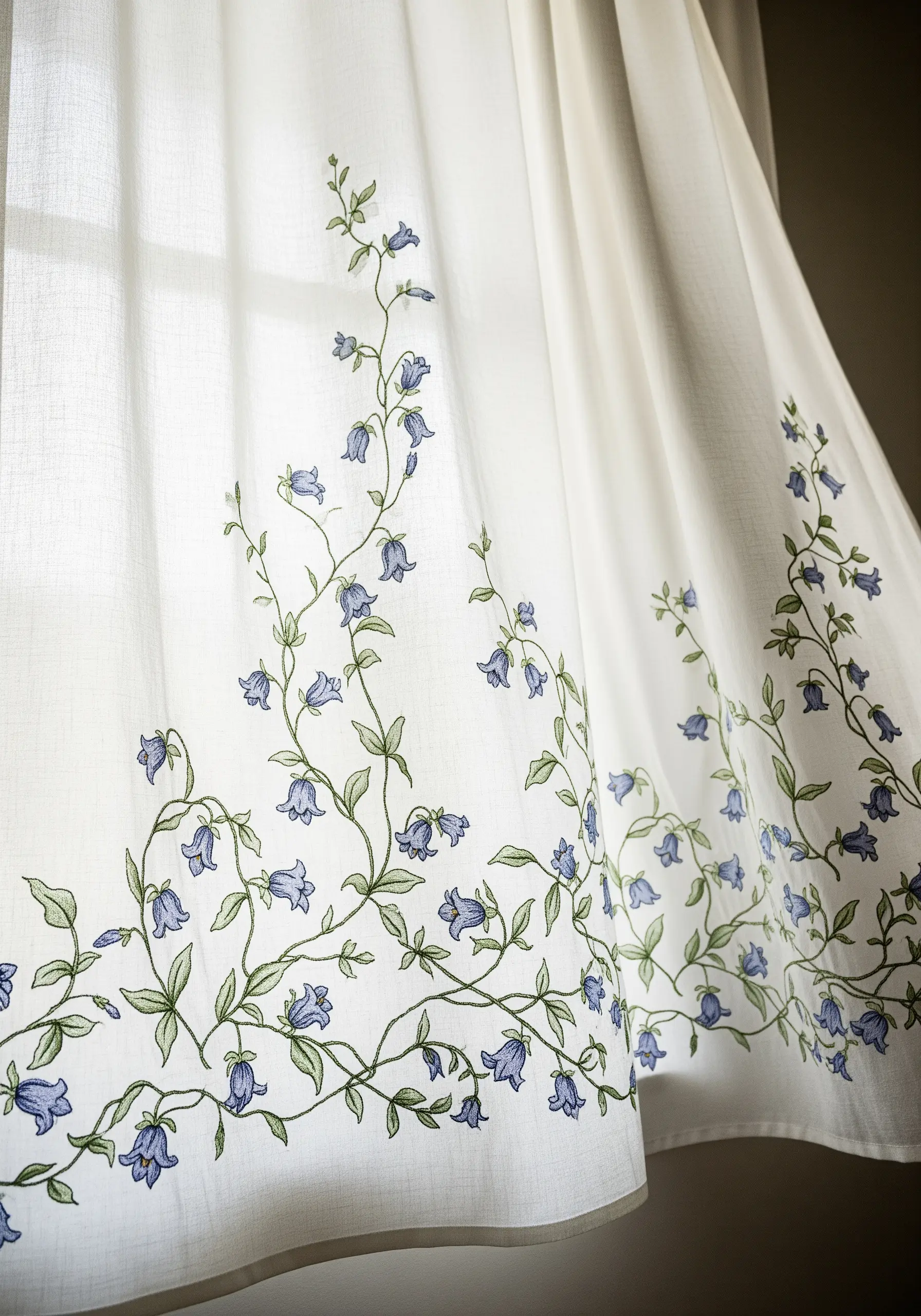 The hem of a sheer white curtain embroidered with a delicate, climbing bluebell vine.