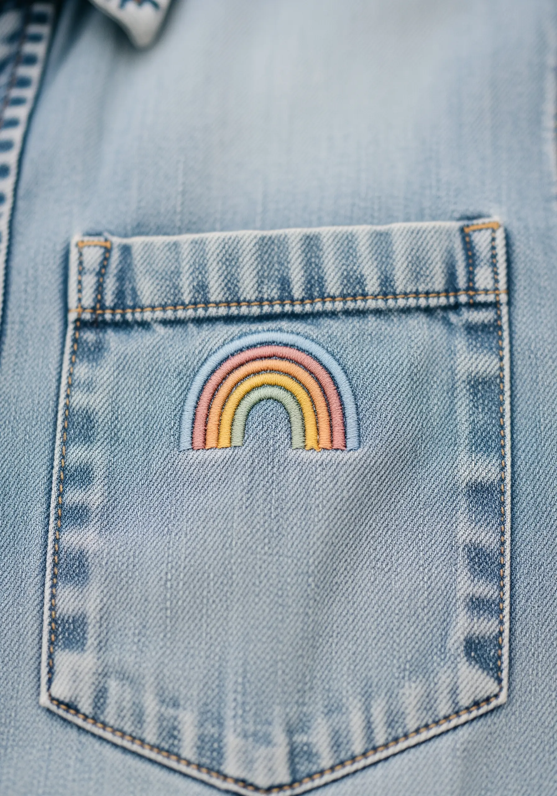 A small, pastel-colored rainbow stitched neatly on the corner of a denim shirt pocket.