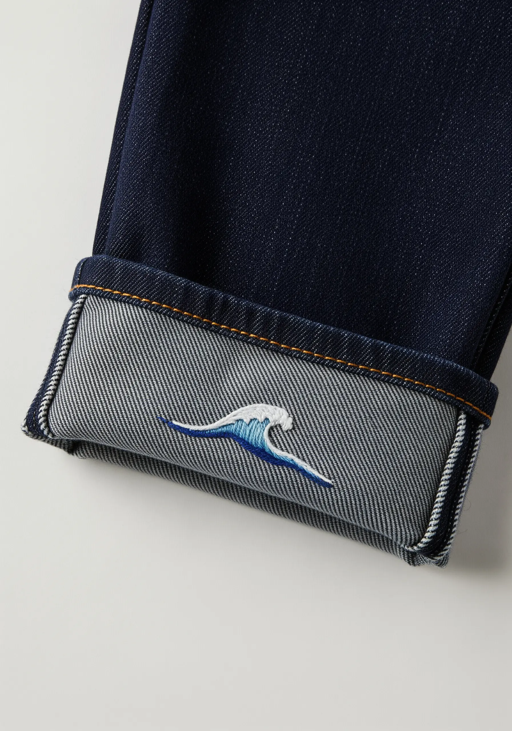 A small, embroidered wave with blue and white gradients on the inside cuff of dark denim jeans.