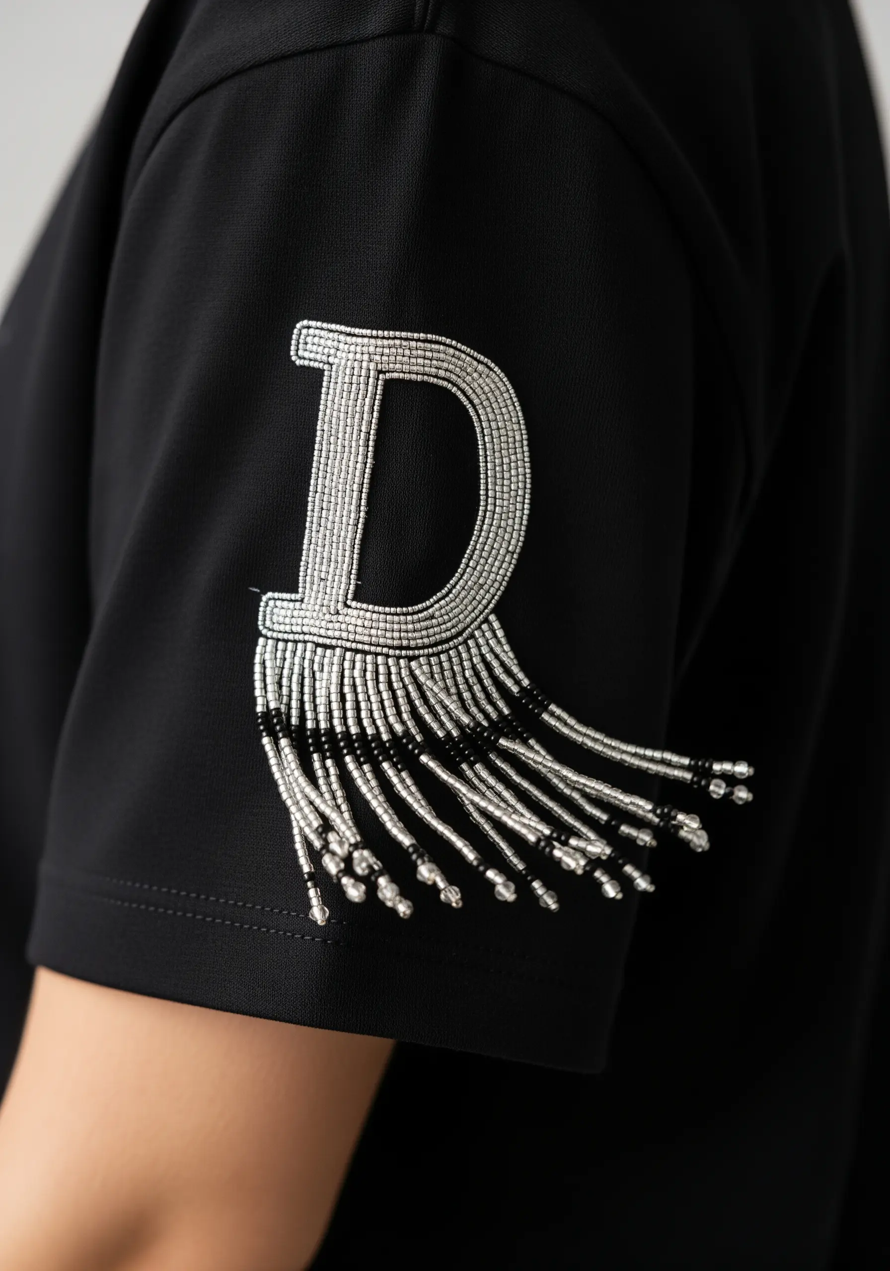 A letter 'D' embroidered with silver beads, featuring a dangling beaded fringe at the bottom.