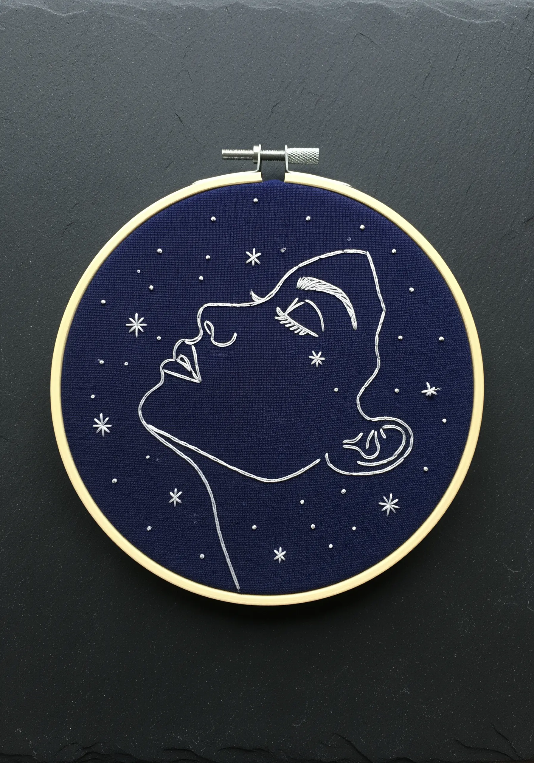 Silver metallic thread line art portrait with small star details on navy blue fabric.