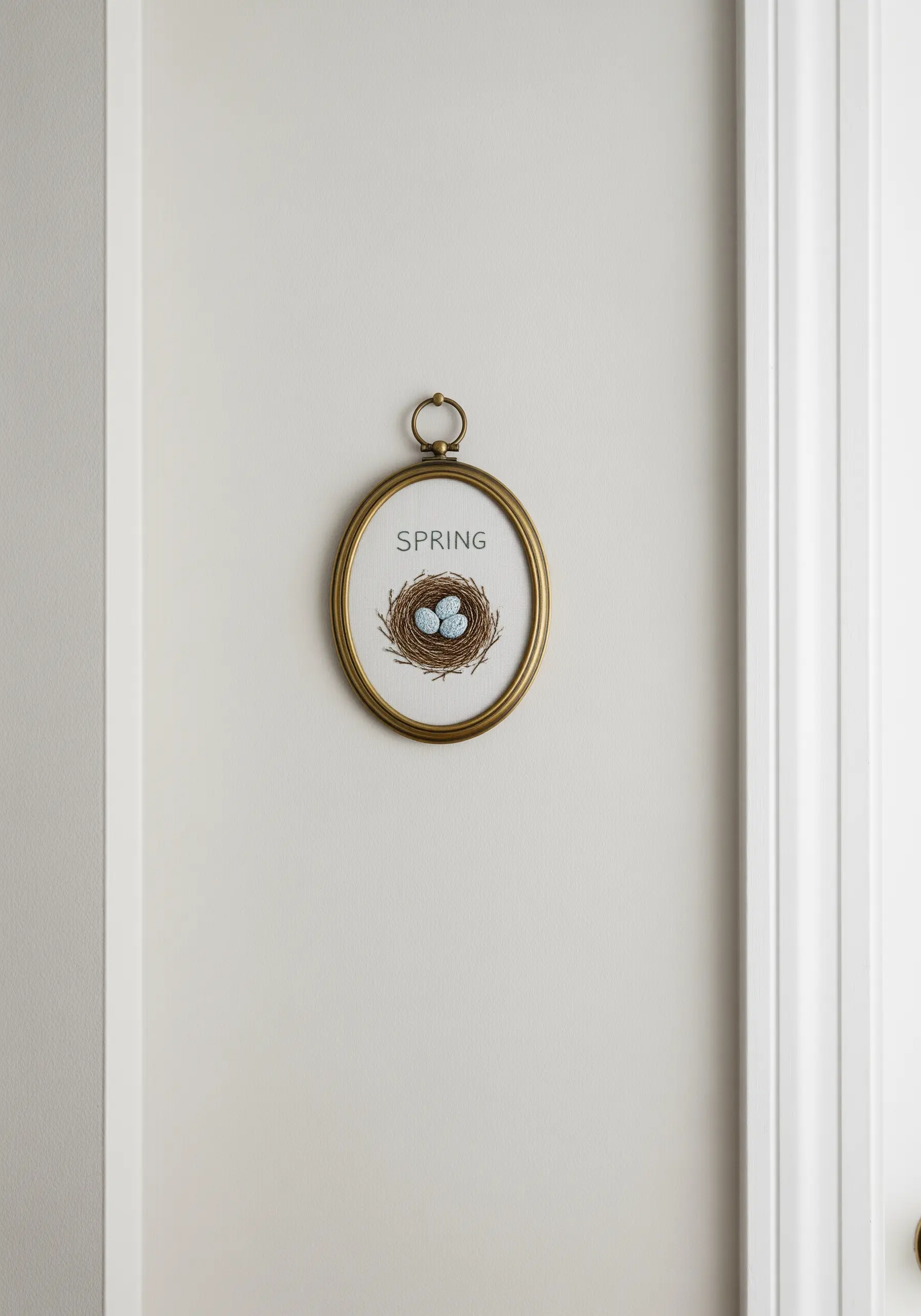 An embroidered bird's nest with blue eggs, framed in a vintage-style oval brass hoop.