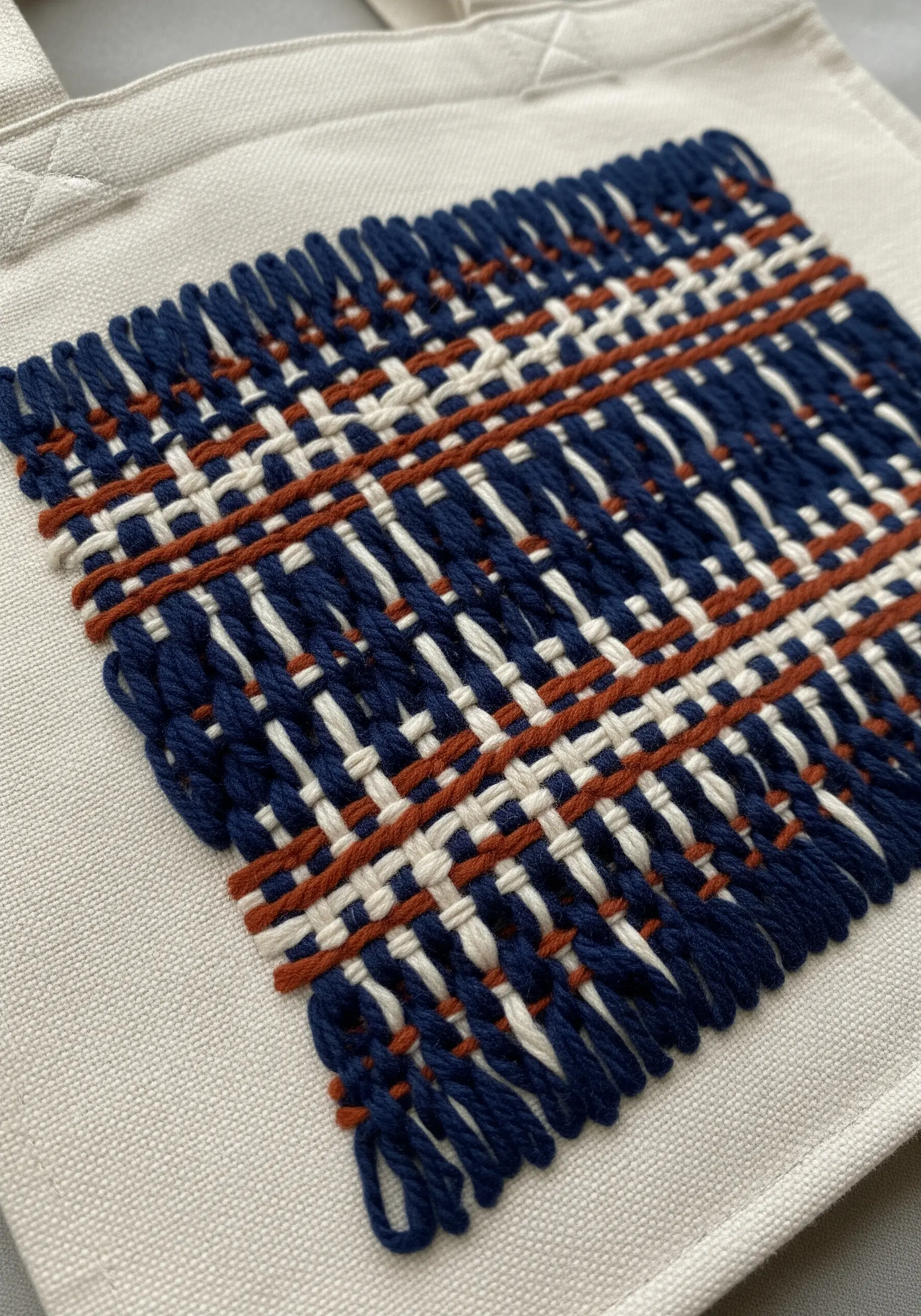 A close-up of a woven patch made with thick blue, white, and rust-colored yarn on a canvas tote.