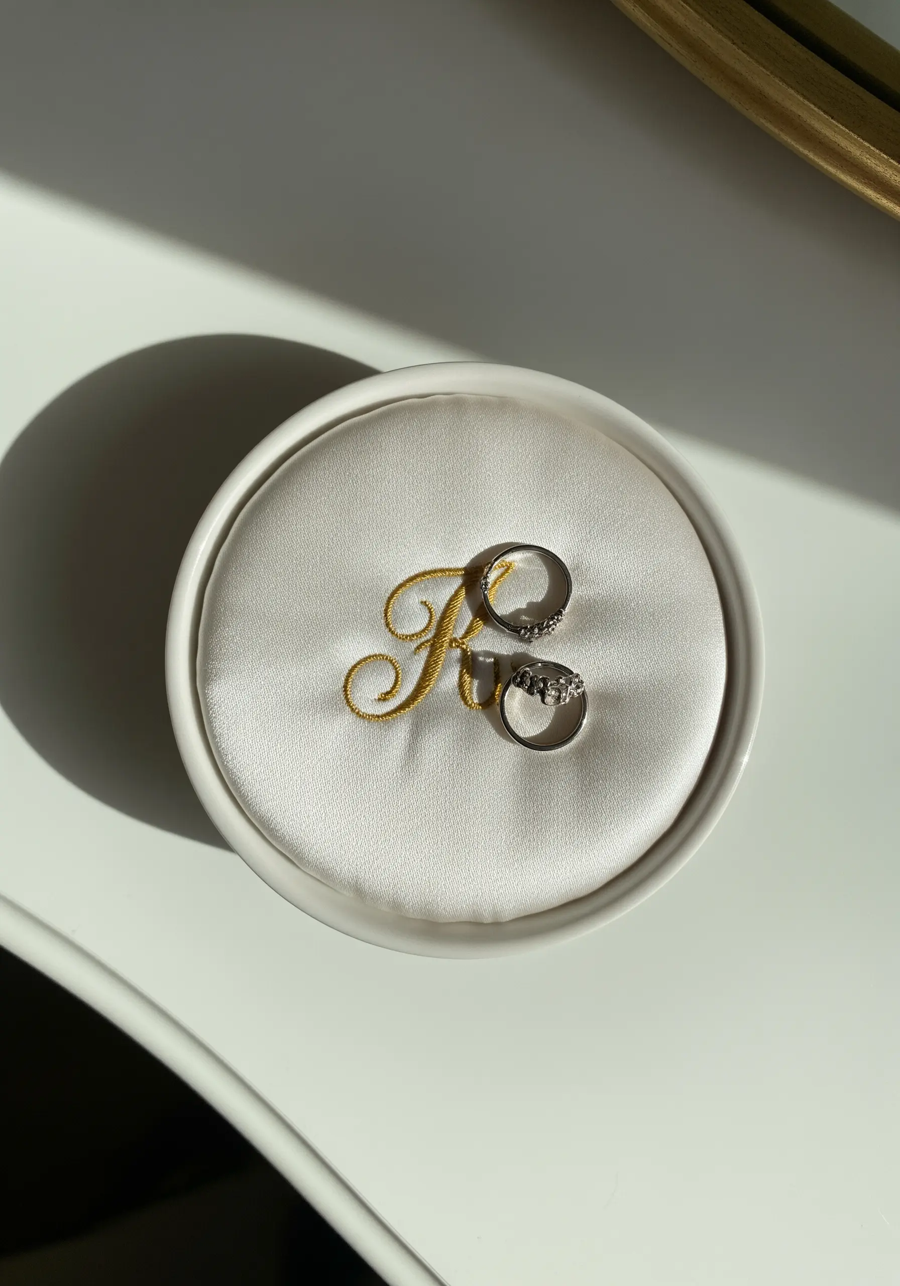 A white ring dish with a gold embroidered letter 'R' on a padded satin surface.