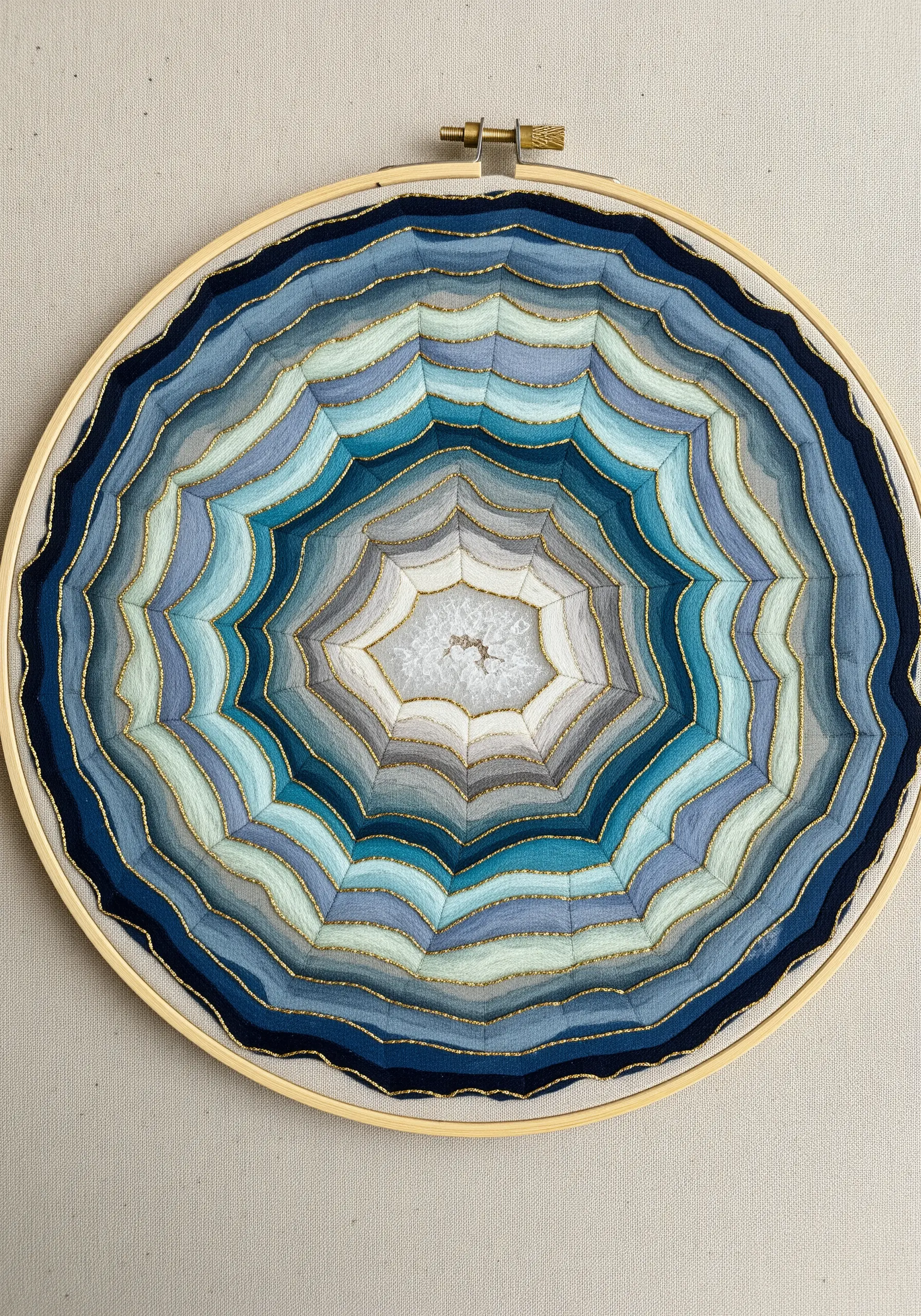 Embroidered geode in a hoop, with concentric rings of blue and grey padded satin stitch.