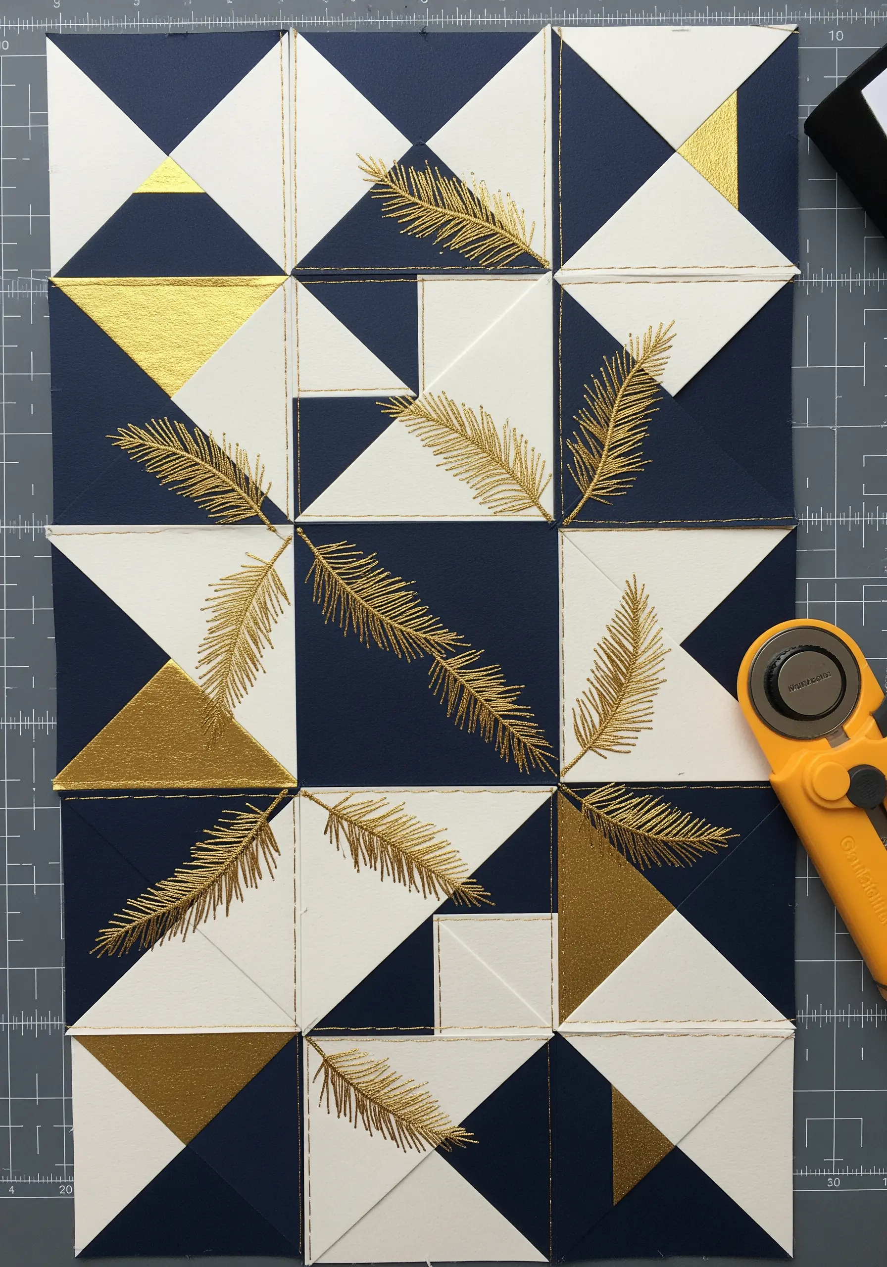 A paper quilt block design with gold metallic thread stitching outlining shapes and forming feathers.