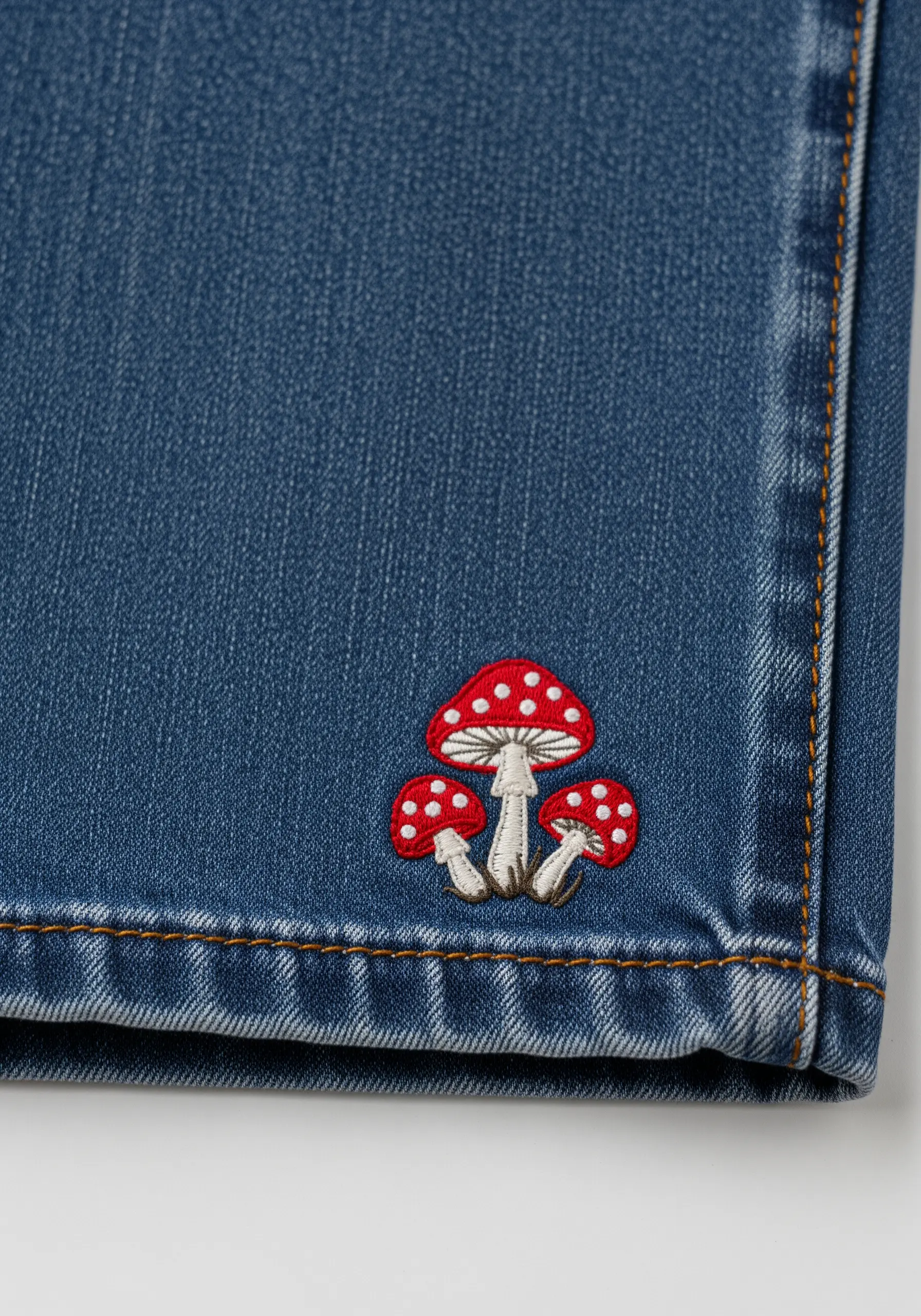 A trio of small red and white embroidered mushrooms on the hem of blue jeans.