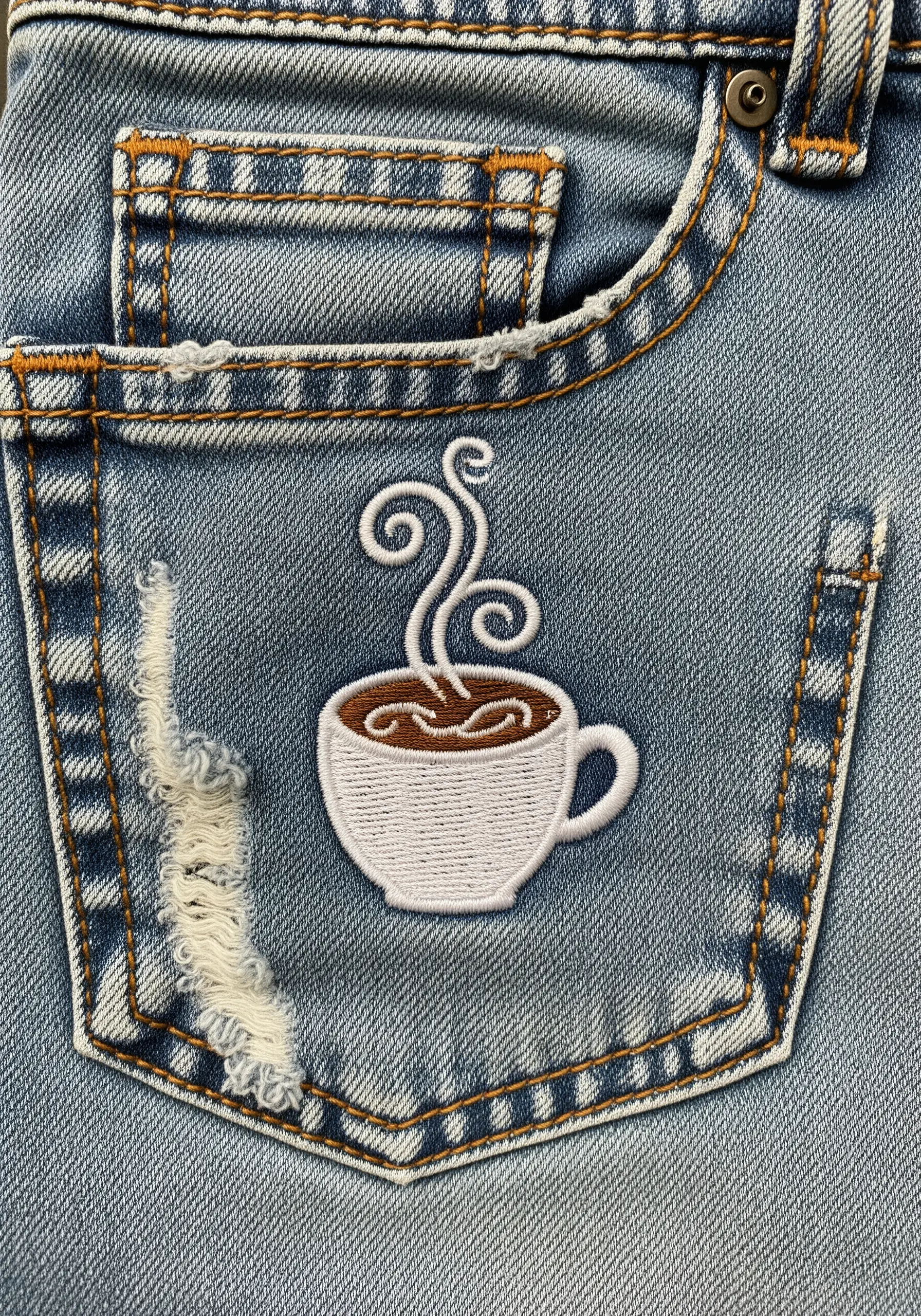 An embroidered coffee cup with whimsical white steam swirls on a distressed denim pocket.