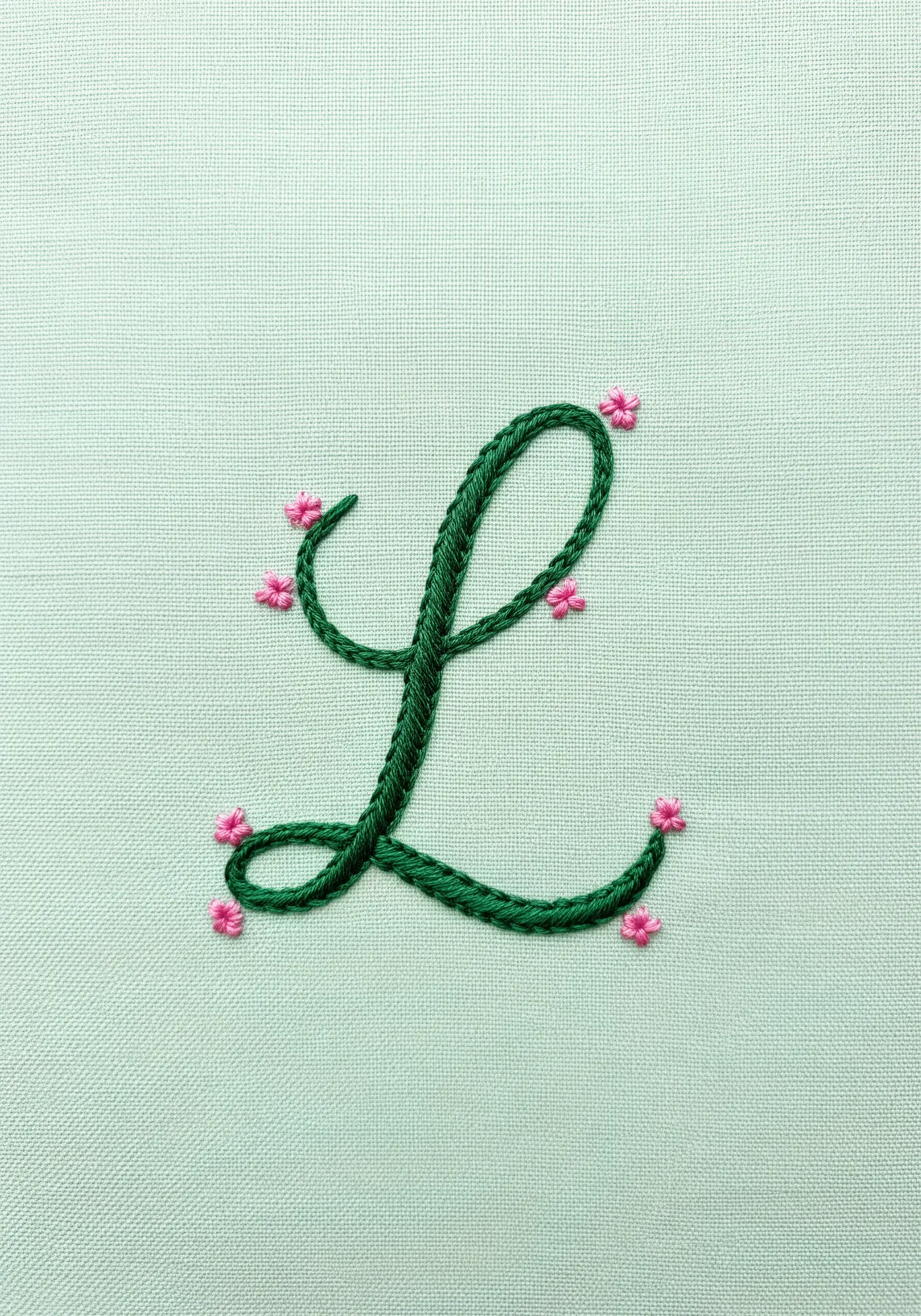 A green cursive letter 'L' decorated with tiny pink lazy daisy flowers.