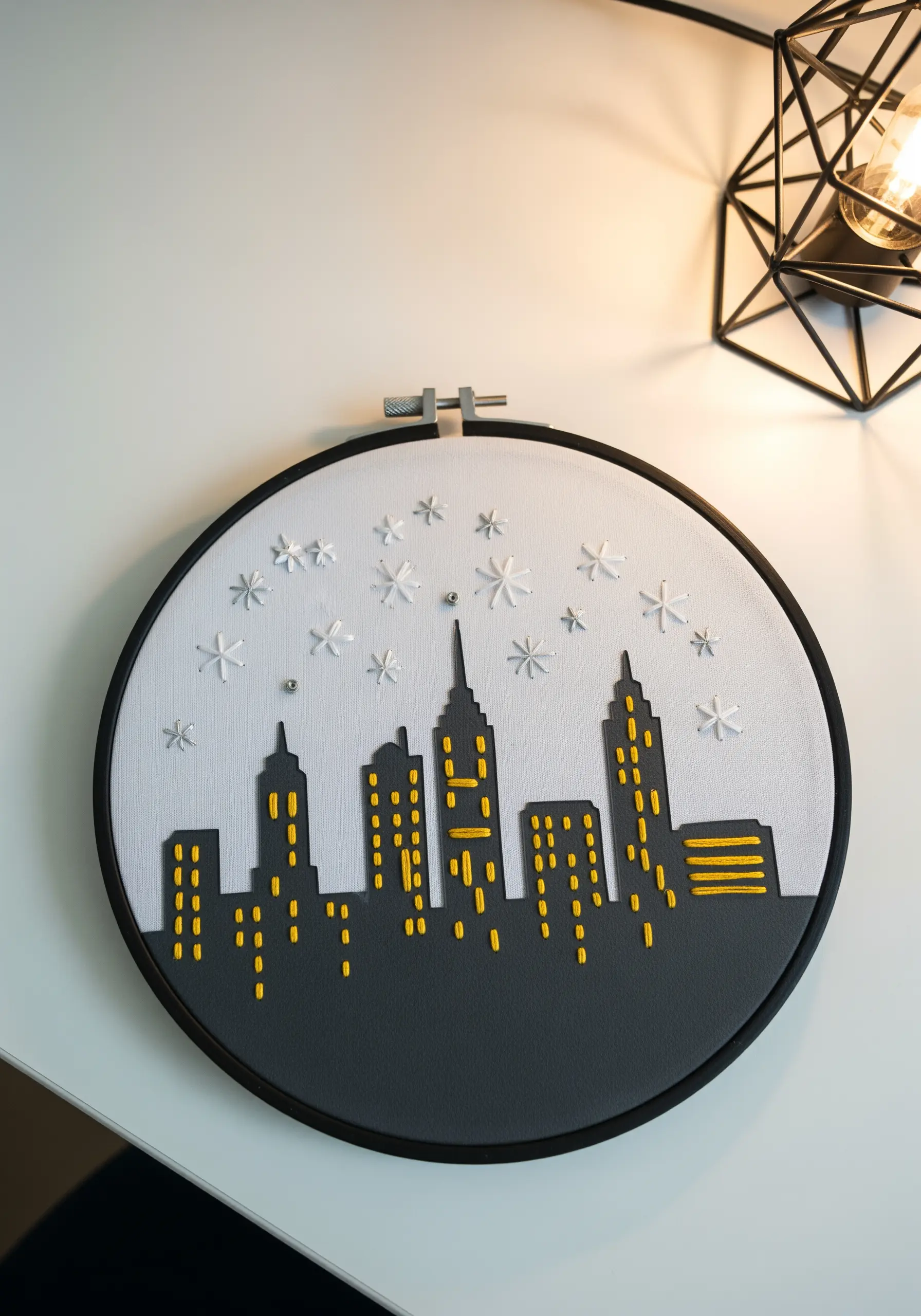 A cityscape hoop art made with paper appliqué buildings and yellow embroidered windows.