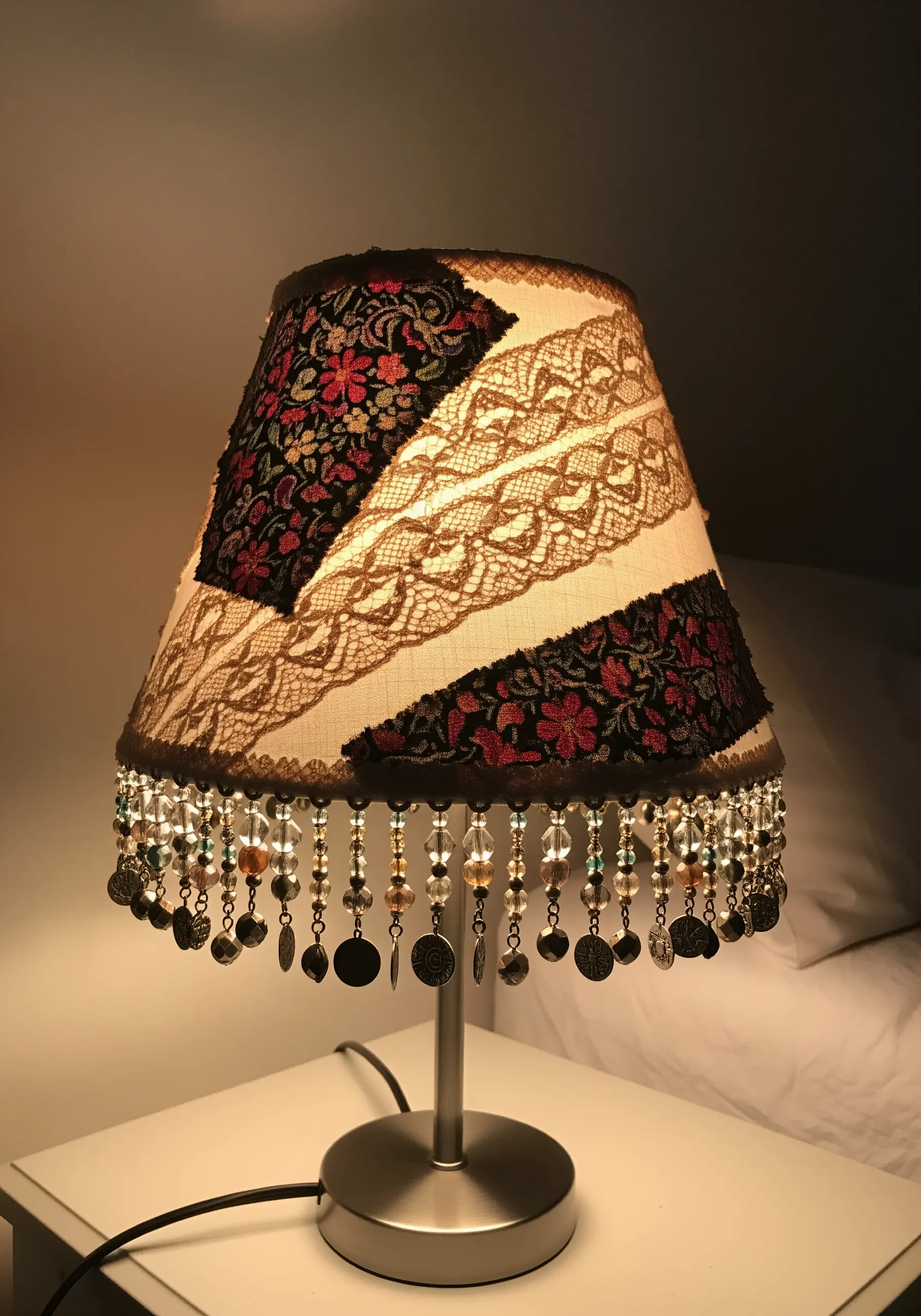 A lampshade decorated with patches of lace, floral fabric, and a beaded fringe.