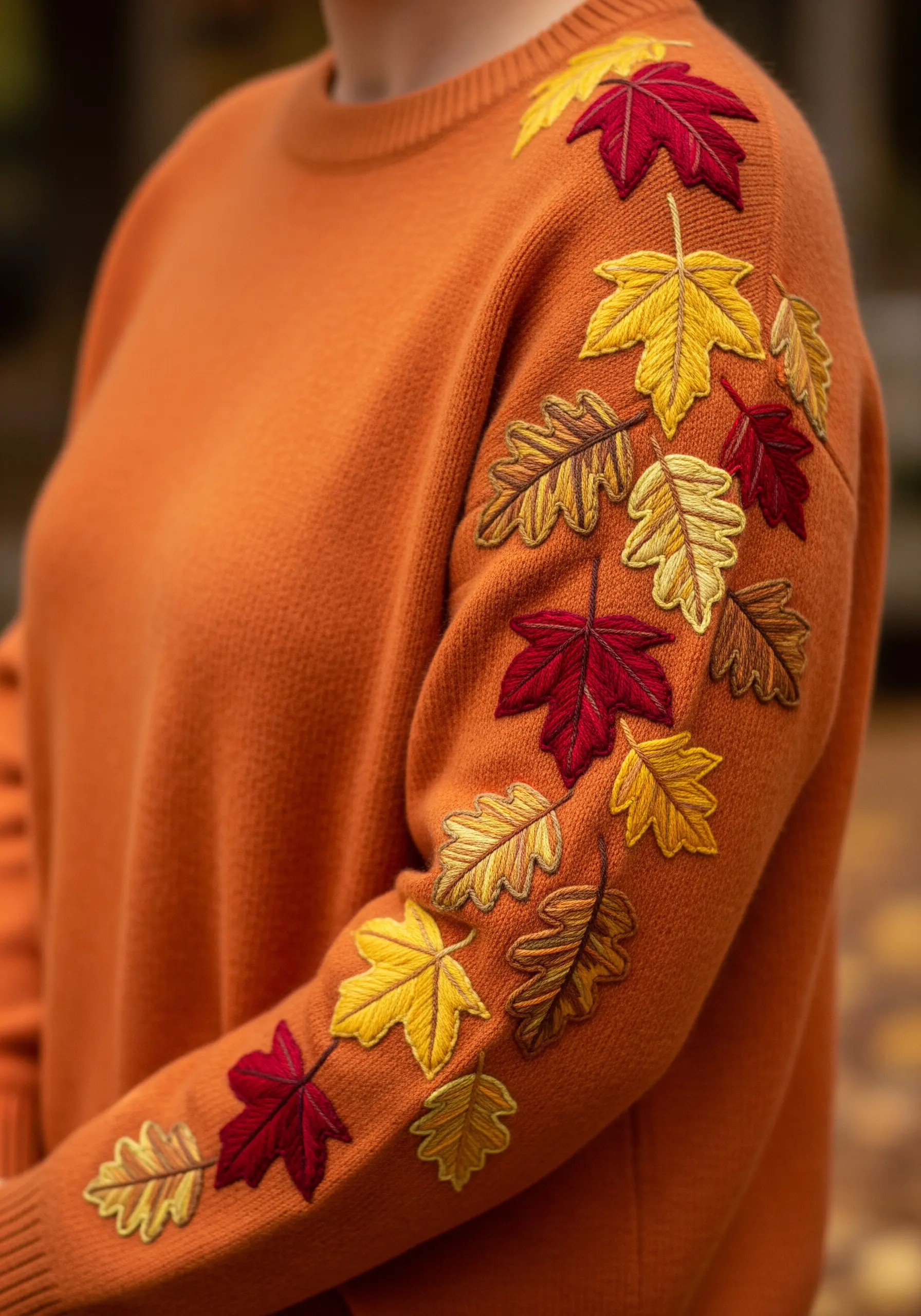 A cascade of embroidered autumn leaves in red, yellow, and orange on an orange knit sweater sleeve.