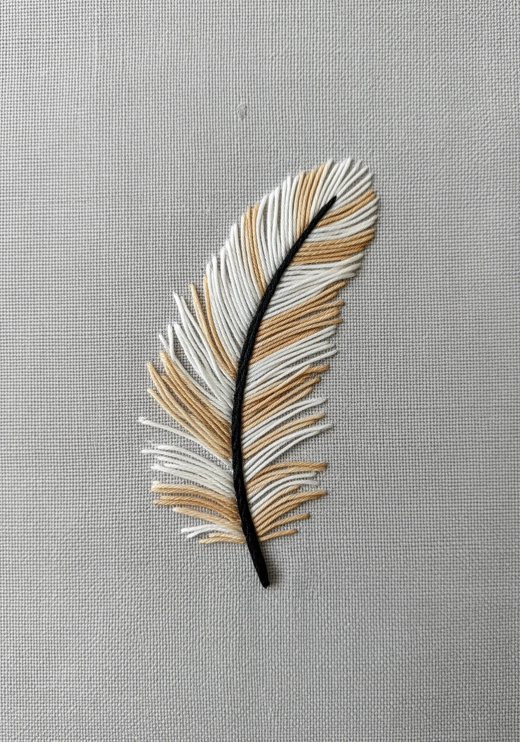 An embroidered feather with blended tan and white threads and a black central quill.
