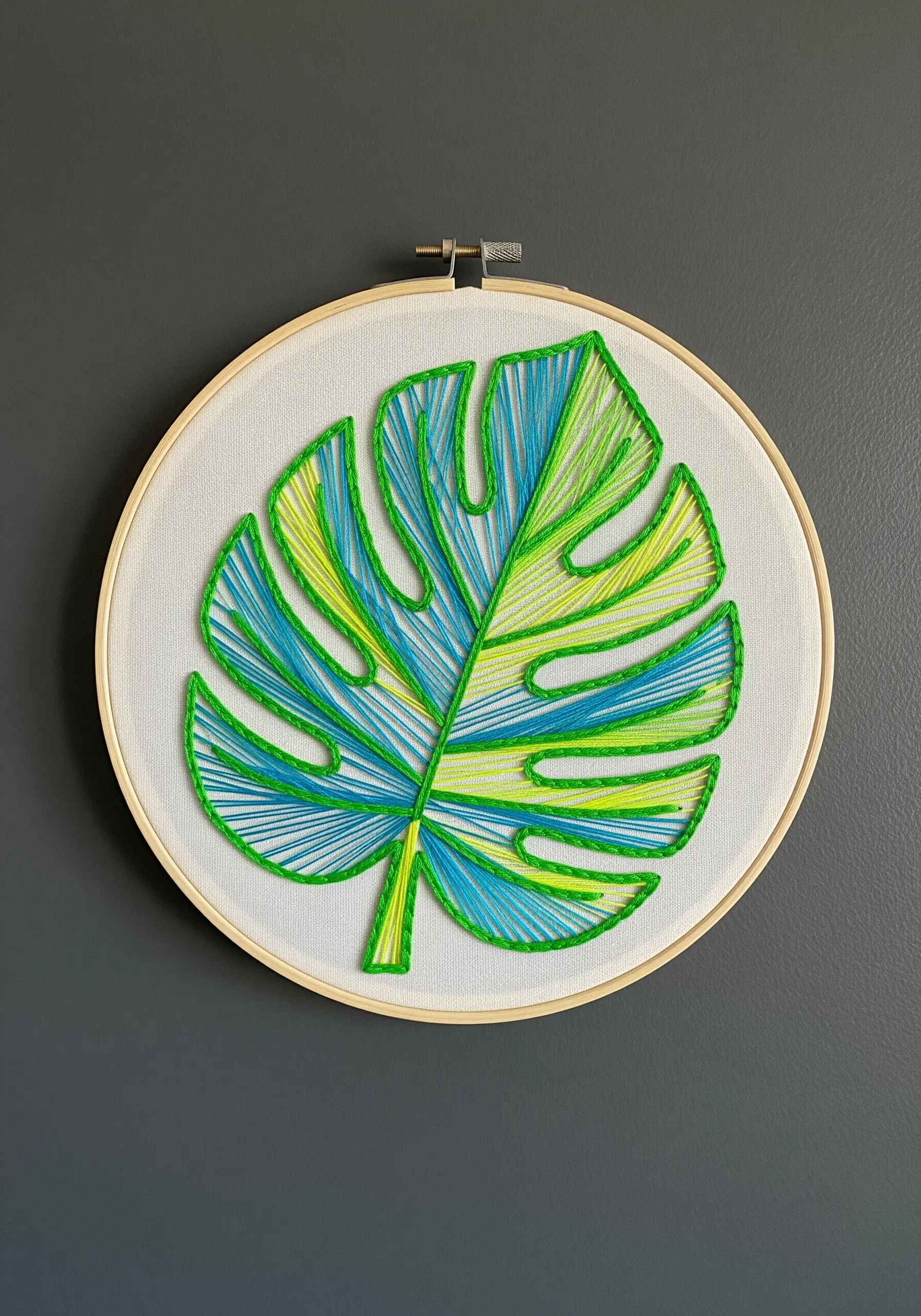 A string art embroidery of a monstera leaf with green, yellow, and blue sections.