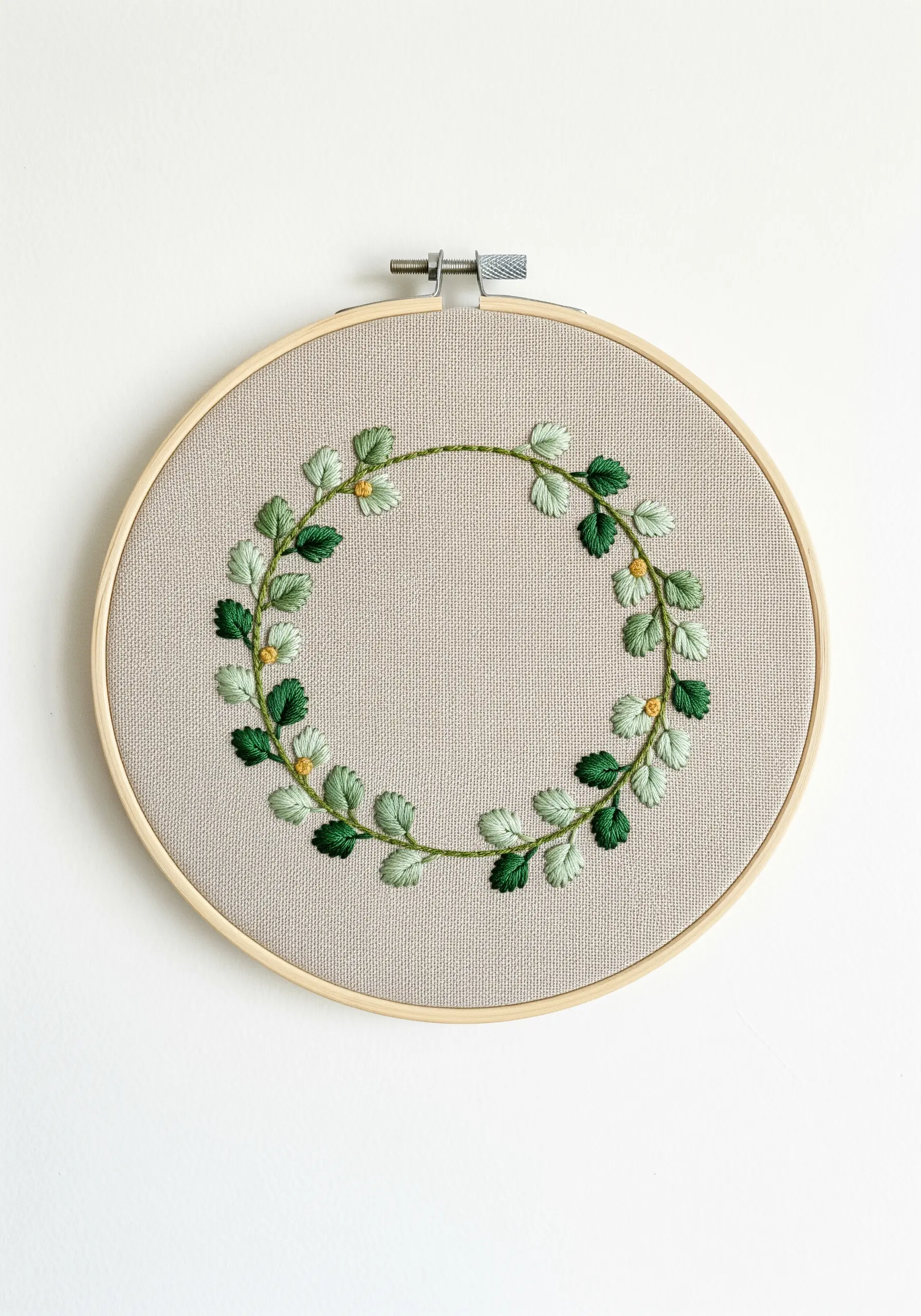 An embroidered wreath of green leaves with small yellow accents in a wooden hoop.