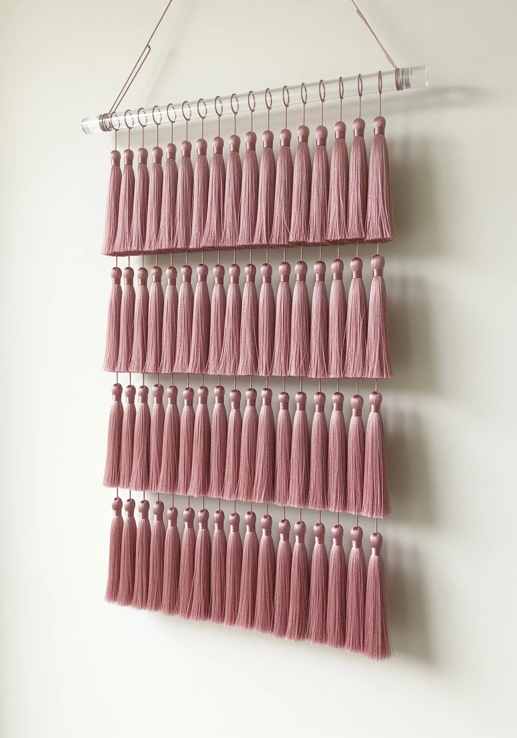 A wall hanging made of four neat rows of dusty pink tassels on a clear acrylic rod.