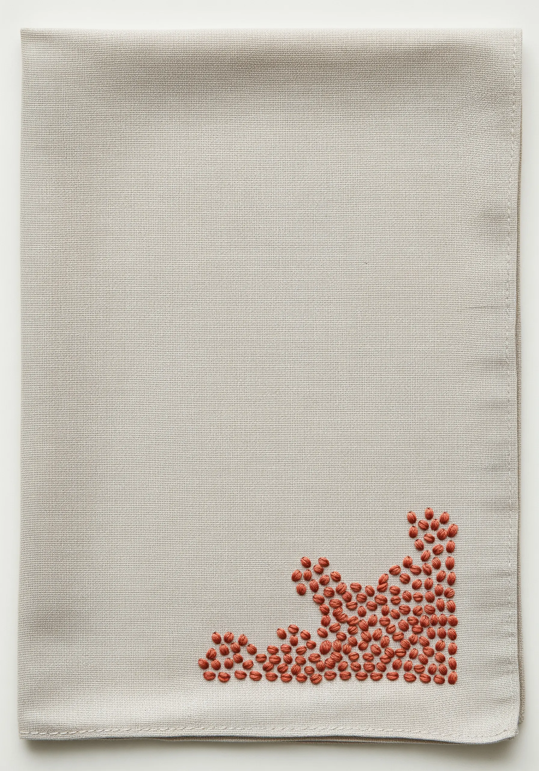 An abstract corner pattern of scattered seed stitches in rust-colored thread on a beige handkerchief.