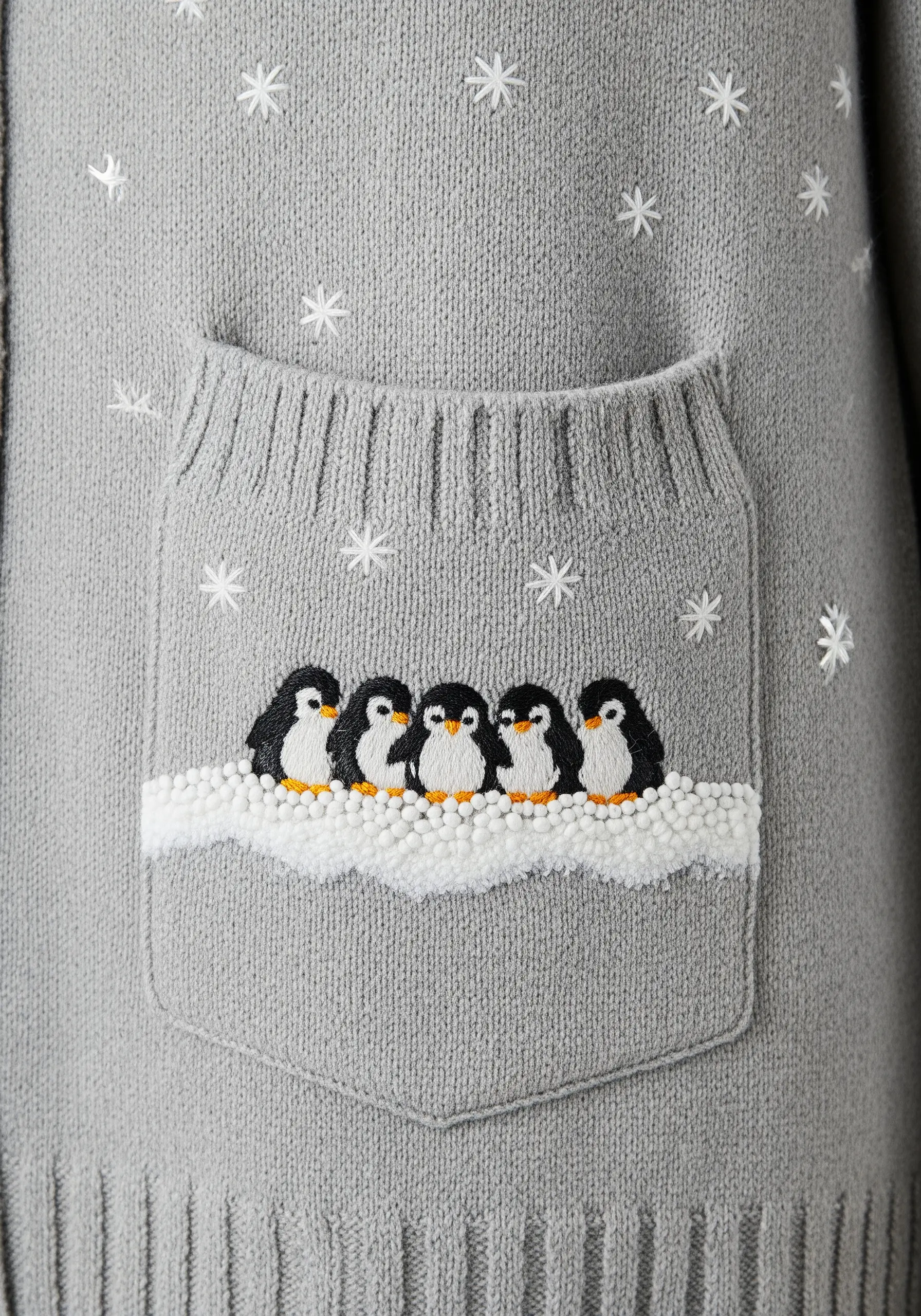A row of small penguins embroidered on a sweater pocket, sitting on textured French knot snow.