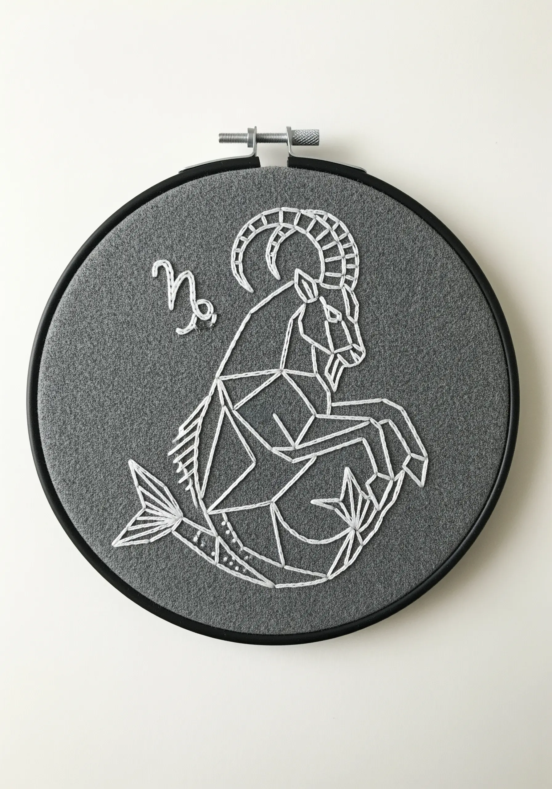 Geometric Capricorn sea-goat embroidered in white on dark grey felt fabric in a black hoop.