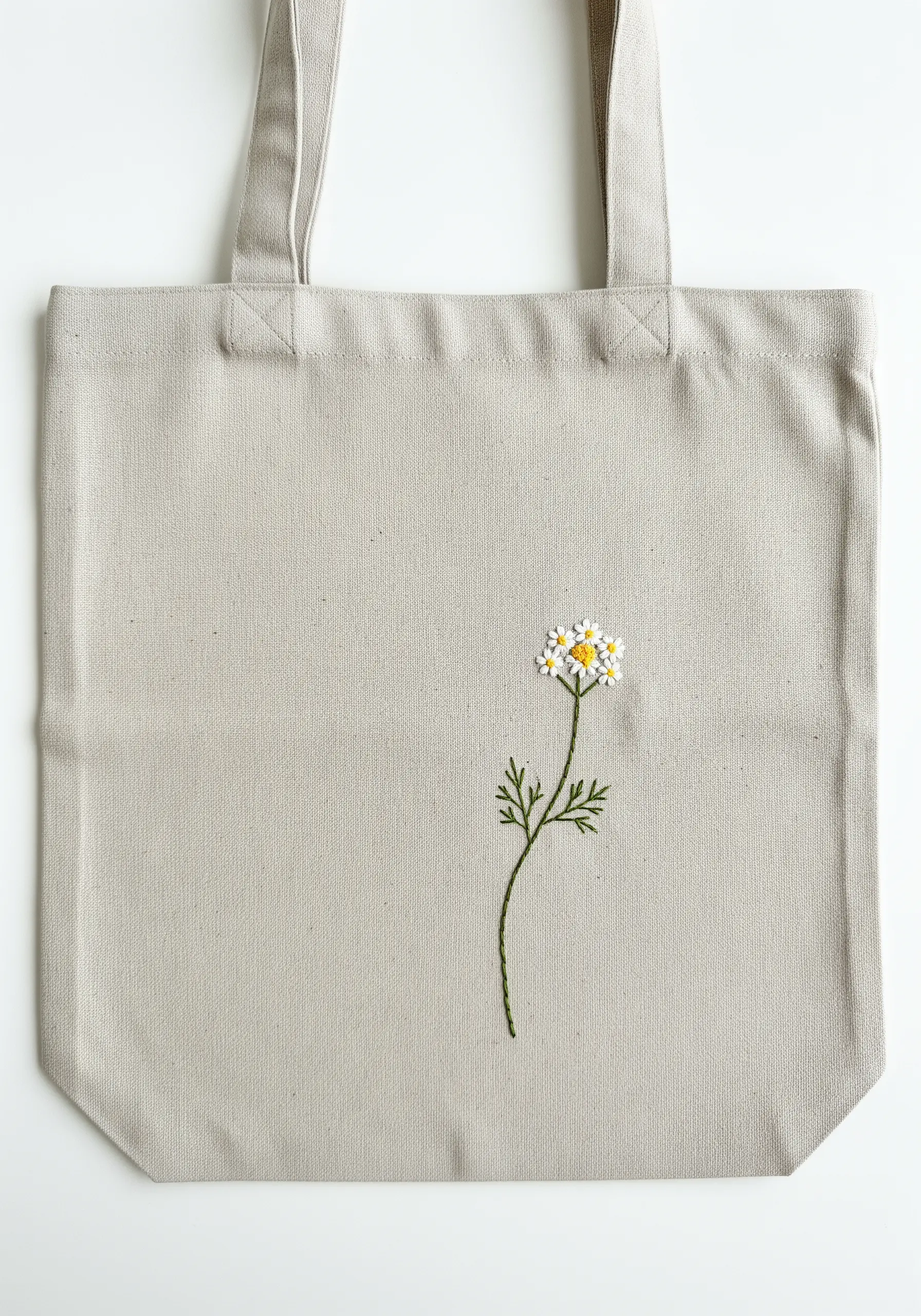 A single embroidered chamomile flower with a green stem on a light grey tote bag.