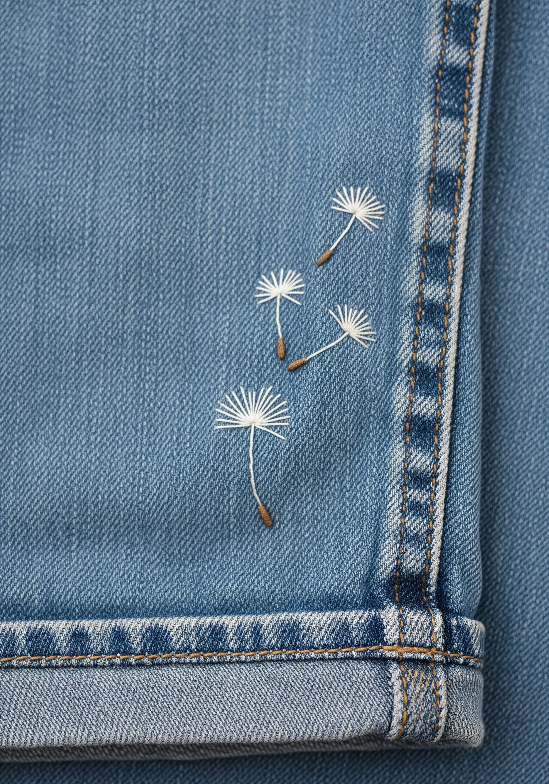 Four delicate dandelion seeds embroidered in white and brown thread, floating up the leg of denim jeans.