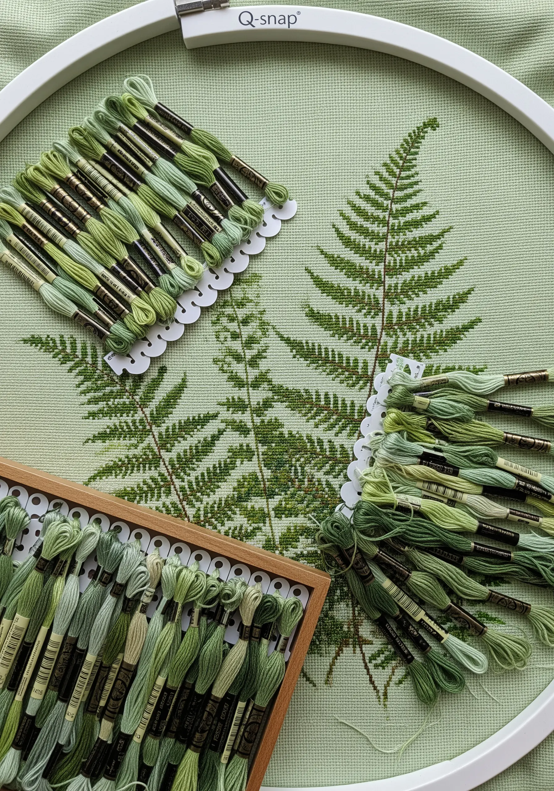 A large cross-stitch project of fern leaves on pale green fabric, with many shades of green thread.