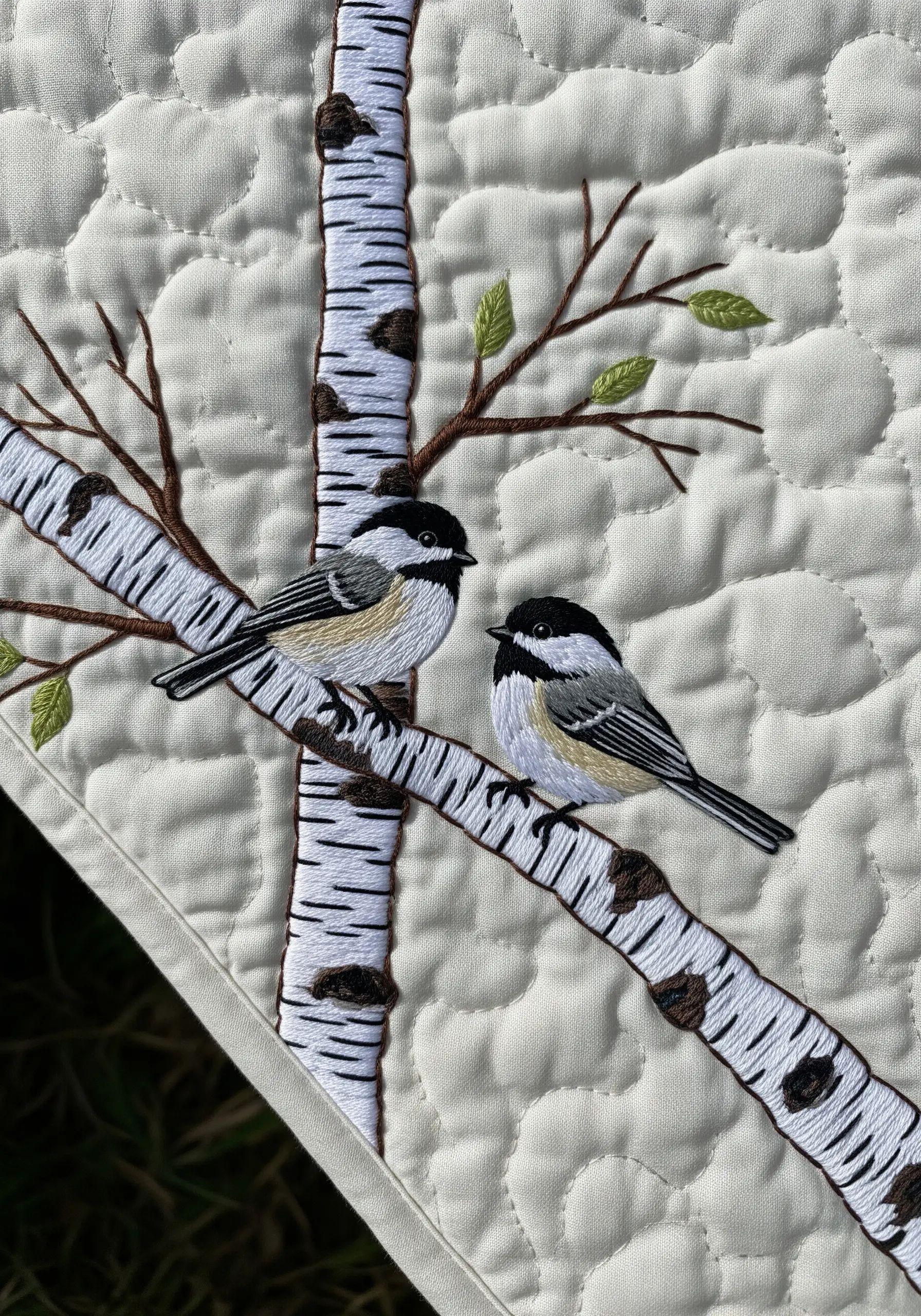 Embroidery of two chickadees perched on a birch tree branch on a quilted blanket.