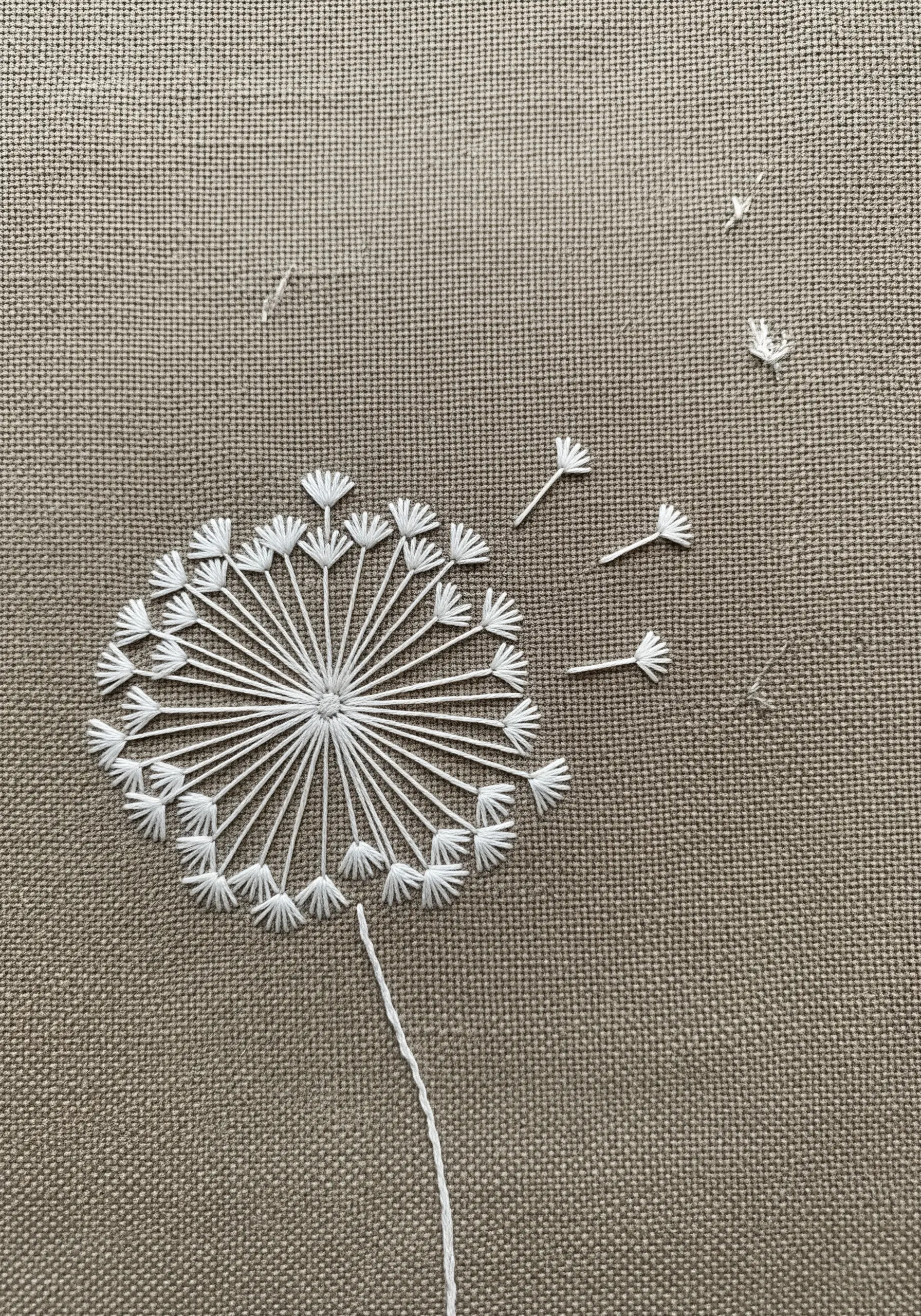 A delicate dandelion seed head with seeds blowing away, embroidered in white on brown linen.