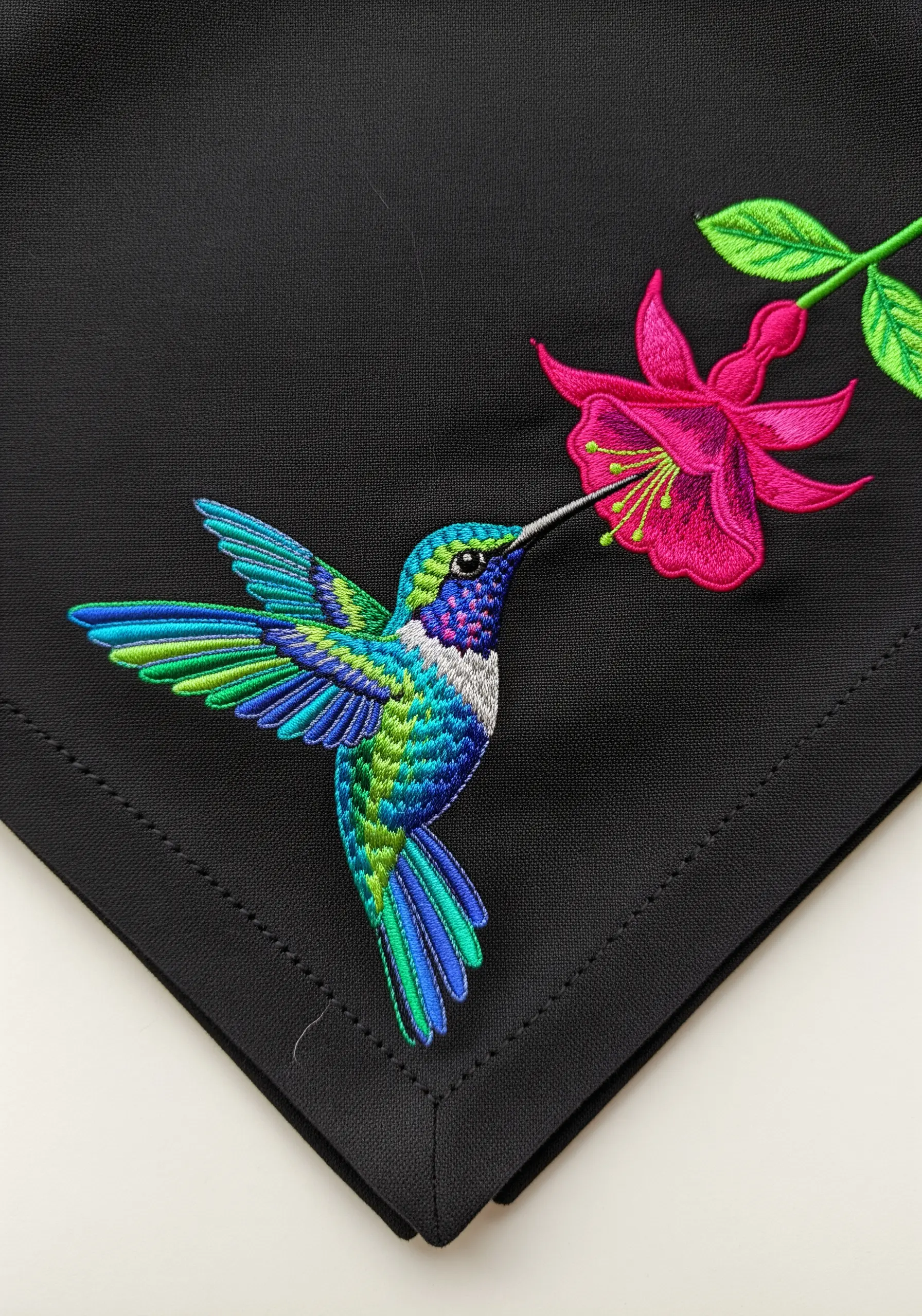 A colorful hummingbird and a pink fuchsia flower embroidered on a black napkin.