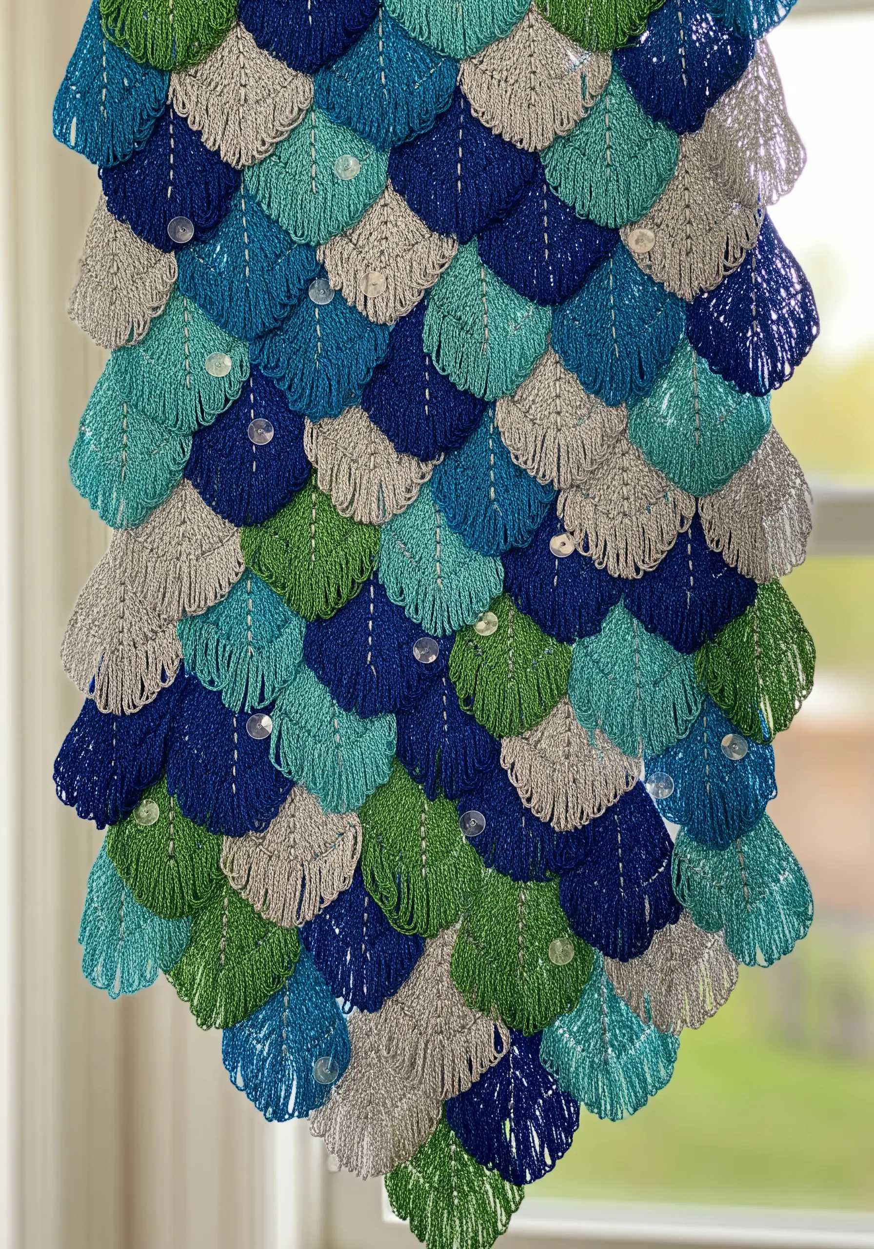 A wall hanging made of layered, scale-like stitches in shades of blue, green, and silver.