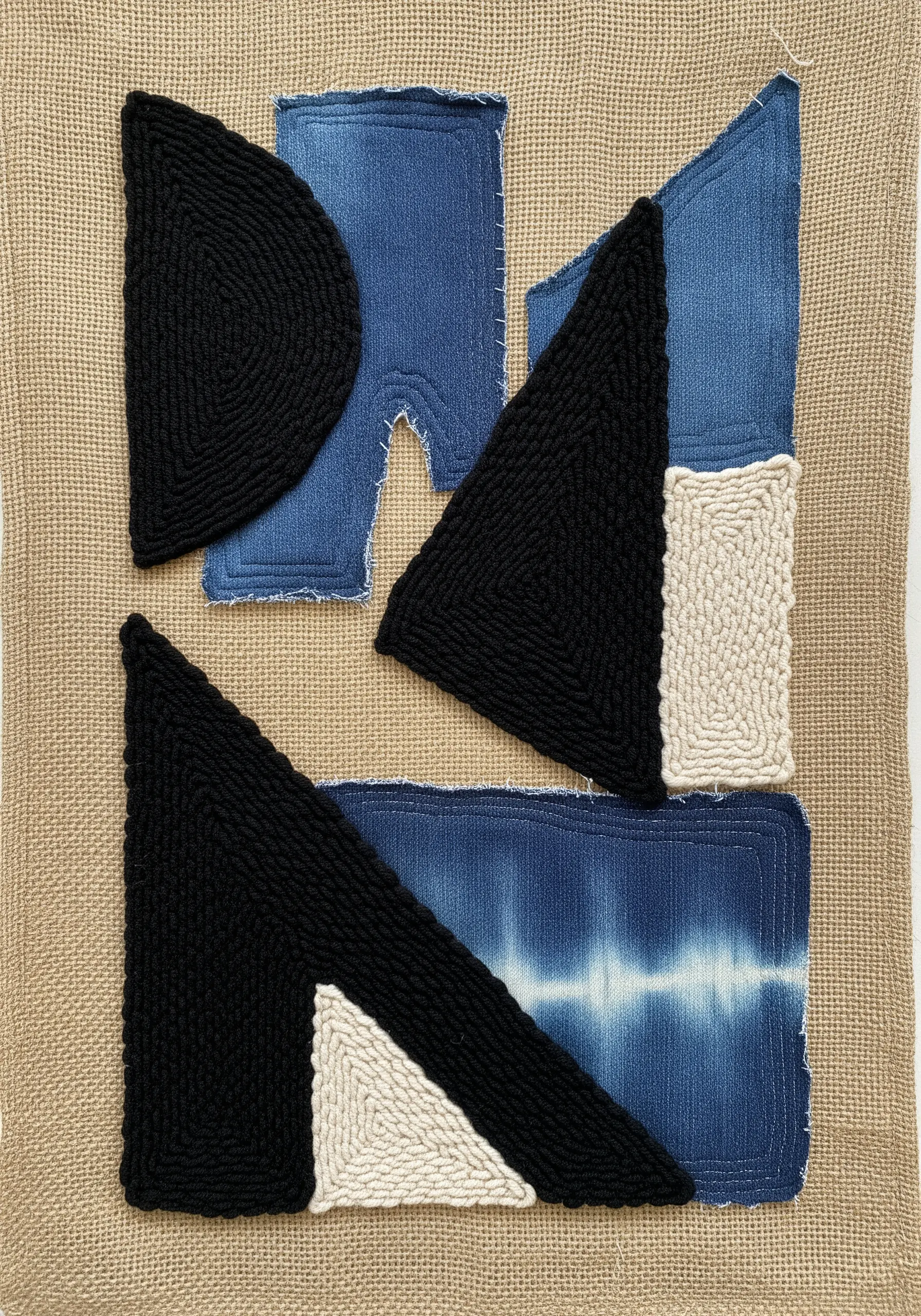 A textile collage on burlap with geometric shapes of denim and black and white punch needle.