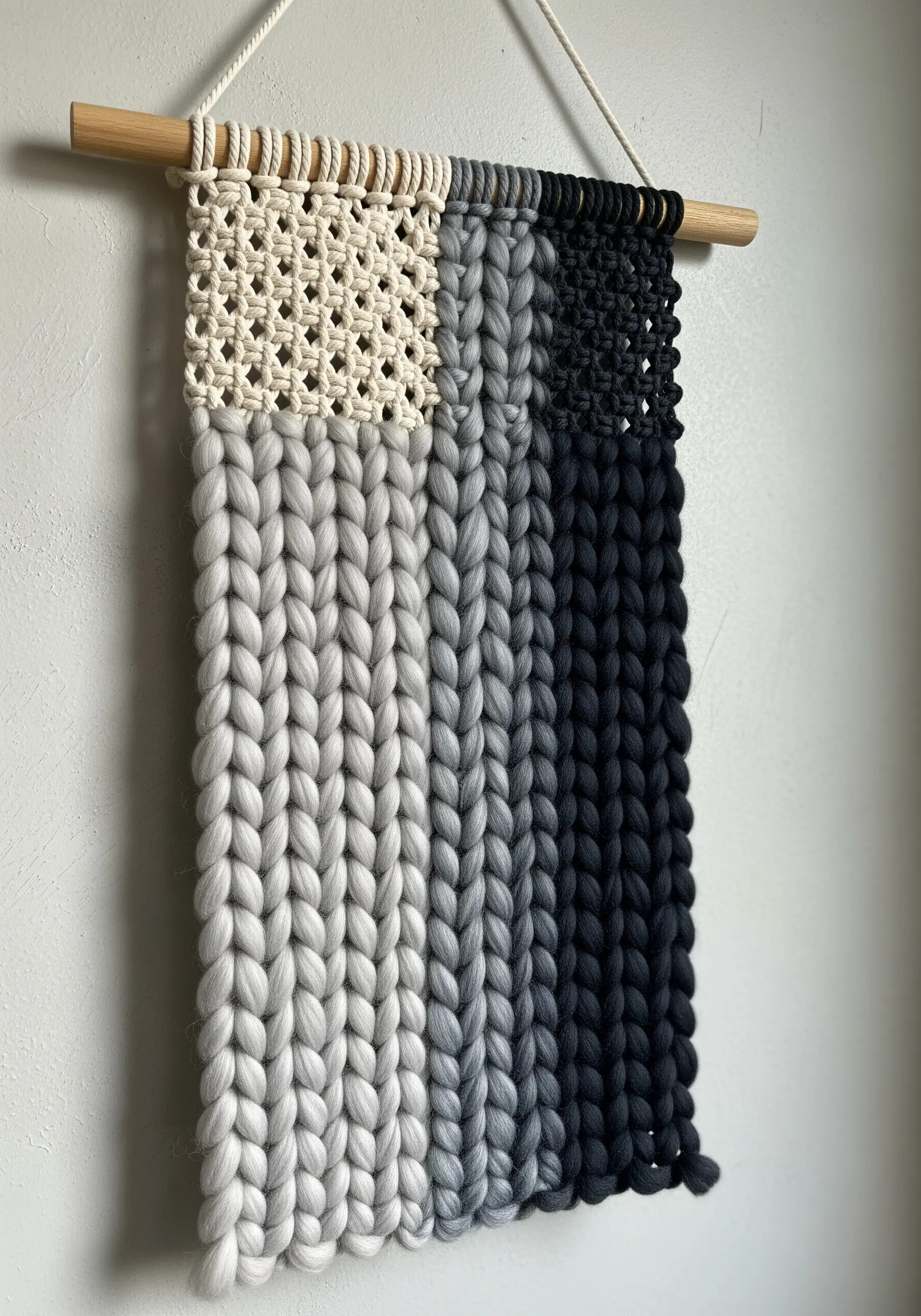 A macrame wall hanging with a color-blocked gradient from black to grey to cream using different knots.