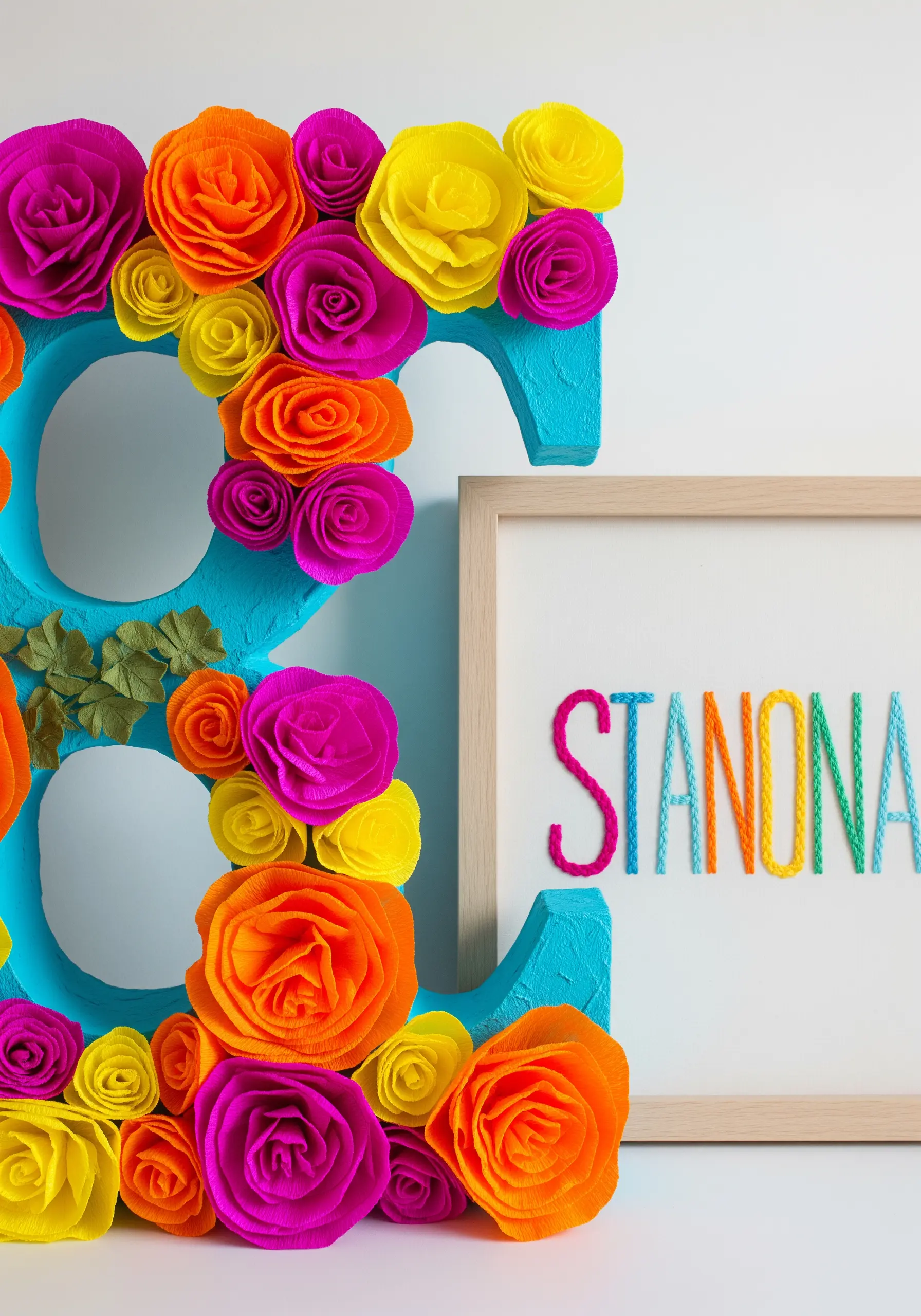 Framed embroidery of a name next to a large letter decorated with colorful crepe paper flowers.