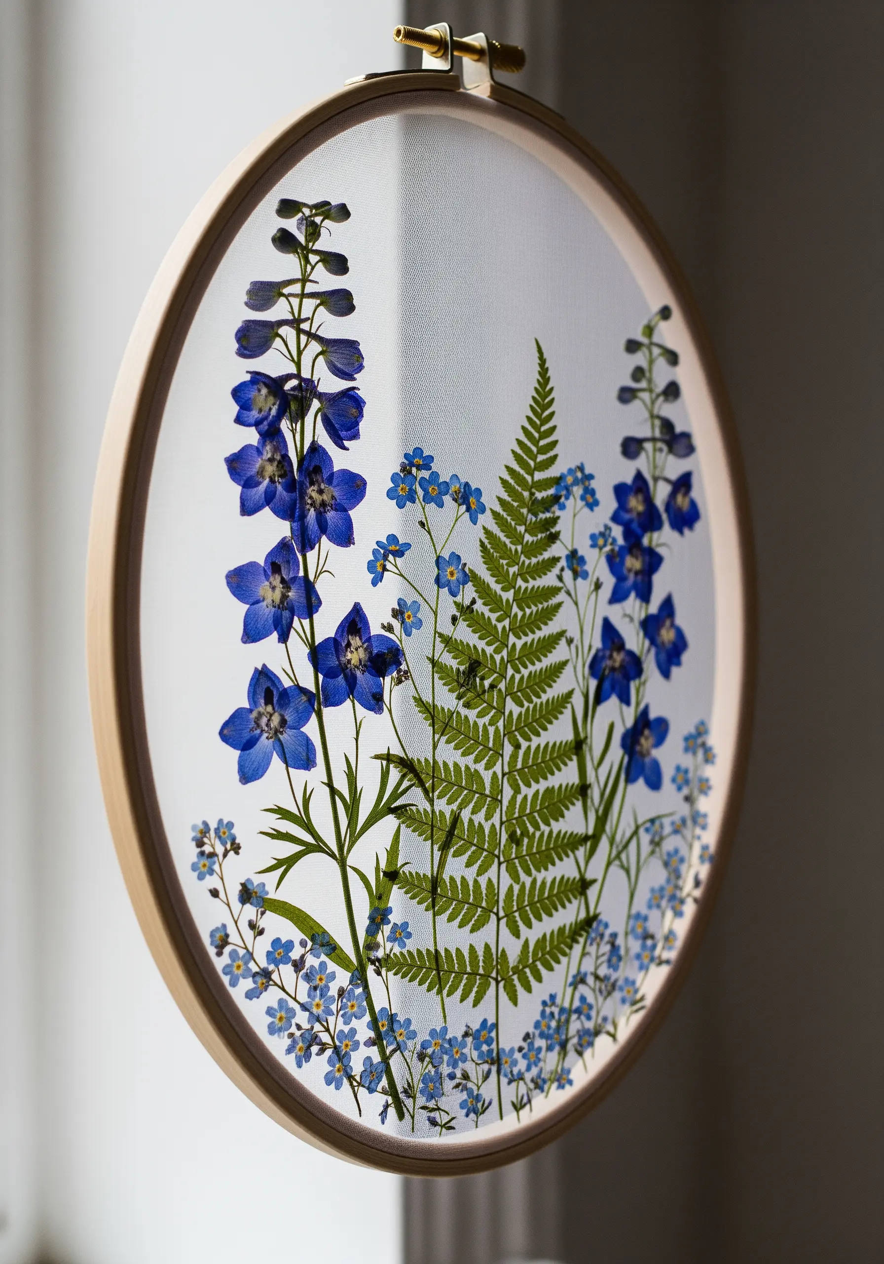 A layered arrangement of pressed blue delphiniums and ferns on sheer fabric in a hoop.