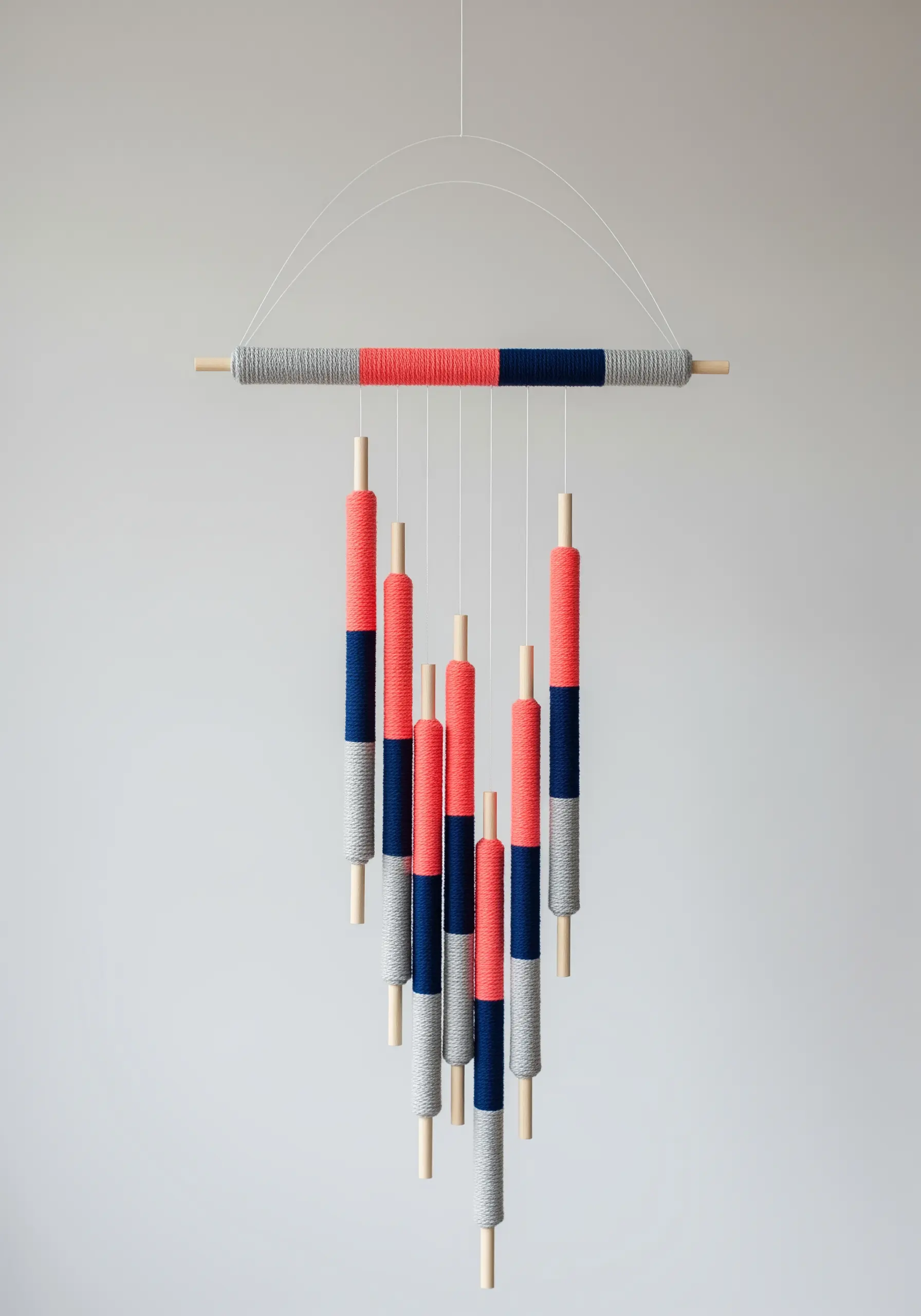 A modern mobile made of yarn-wrapped dowels in coral, navy, and grey, hanging from a larger dowel.